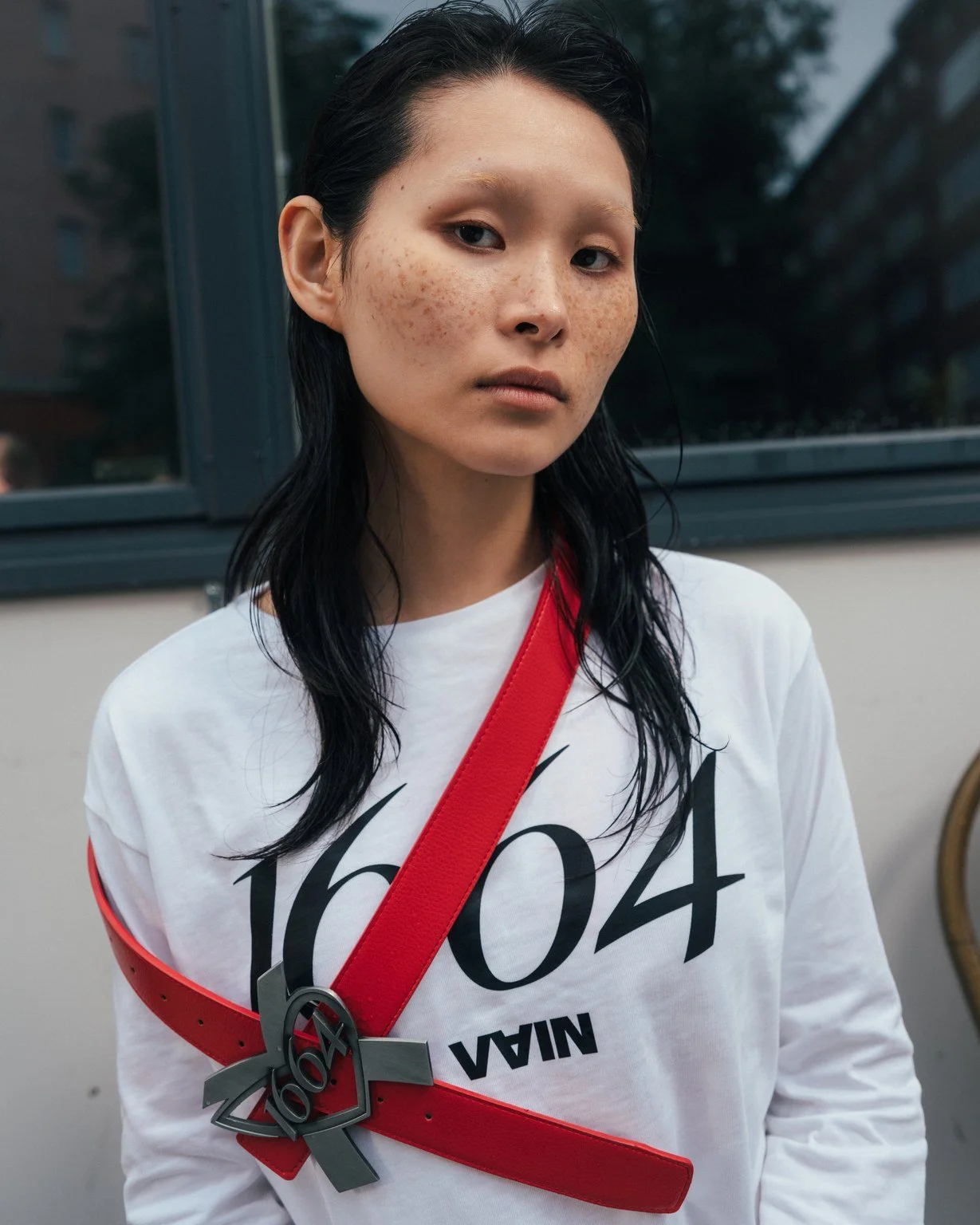 Young woman with long black hair and freckles wearing a white shirt with black text, a red medal ribbon, and a silver Olympic medal around her neck, standing indoors near a window.