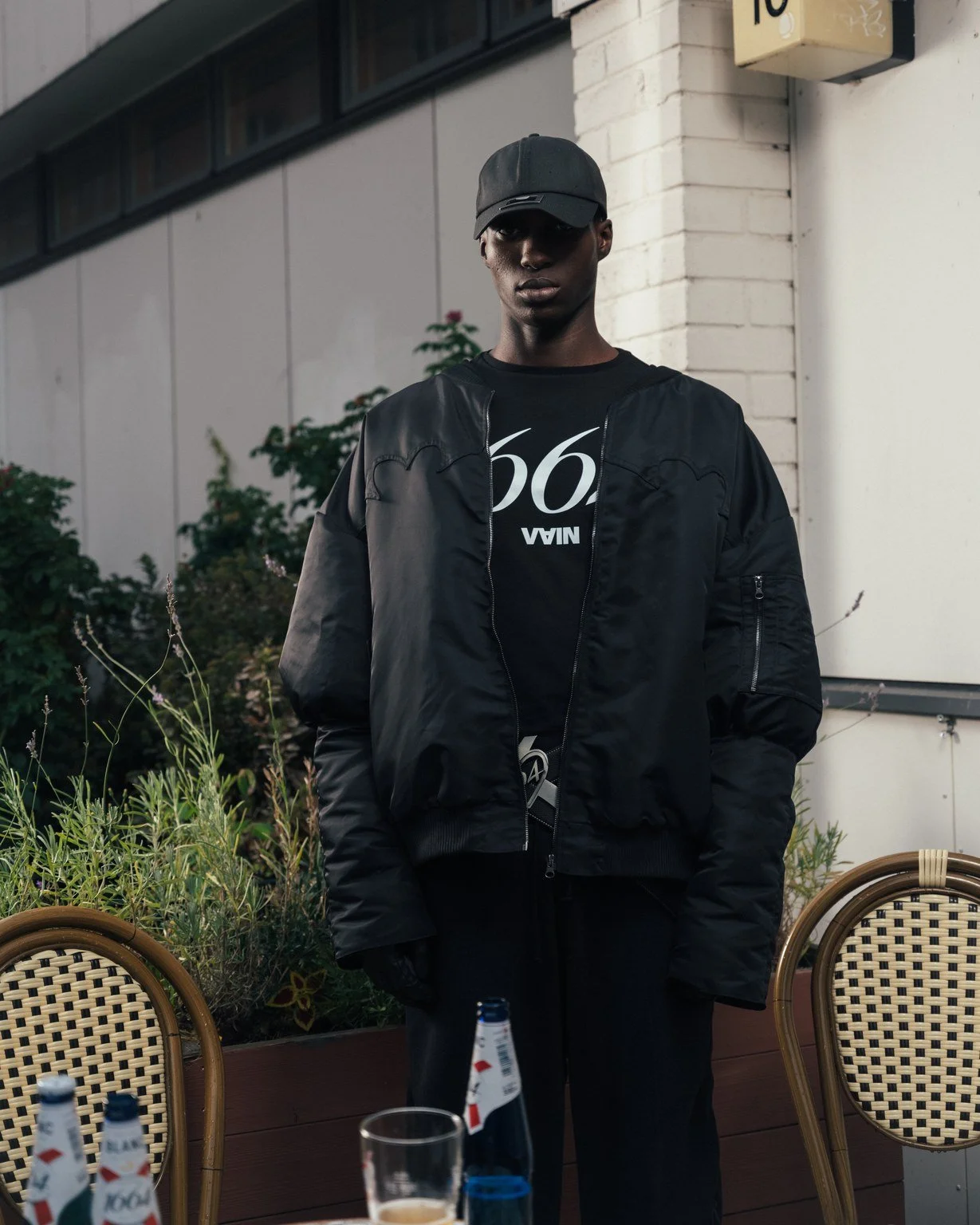 A young man wearing a black cap, black jacket, and black gloves stands outdoors near a white brick wall and some plants. There are chairs and bottles on the table in front of him.