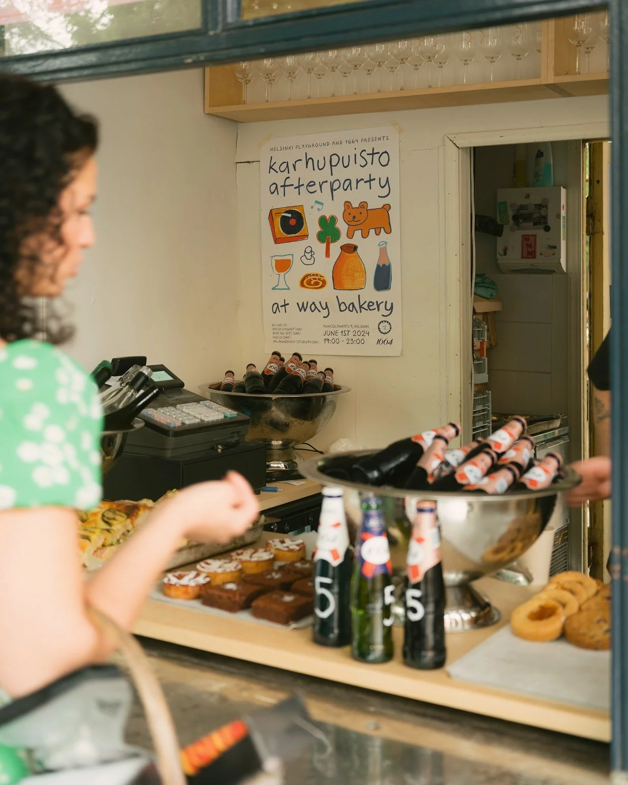 A bakery counter with various baked goods and bottles, with a woman in a green dress on the left side. There is a poster on the wall behind the counter that reads 'Karhupuisto afterparty at way bakery' with colorful illustrations of a bear, a beer, a