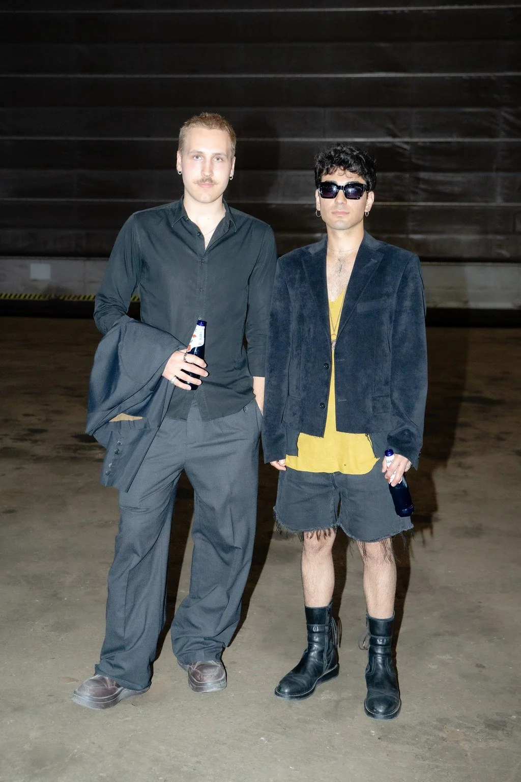 Two men standing in a dark, industrial setting, each holding a beer bottle. The man on the left wears a black button-up shirt and gray pants, with a blazer over his arm. The man on the right wears sunglasses, a dark jacket, a yellow shirt, ripped sho