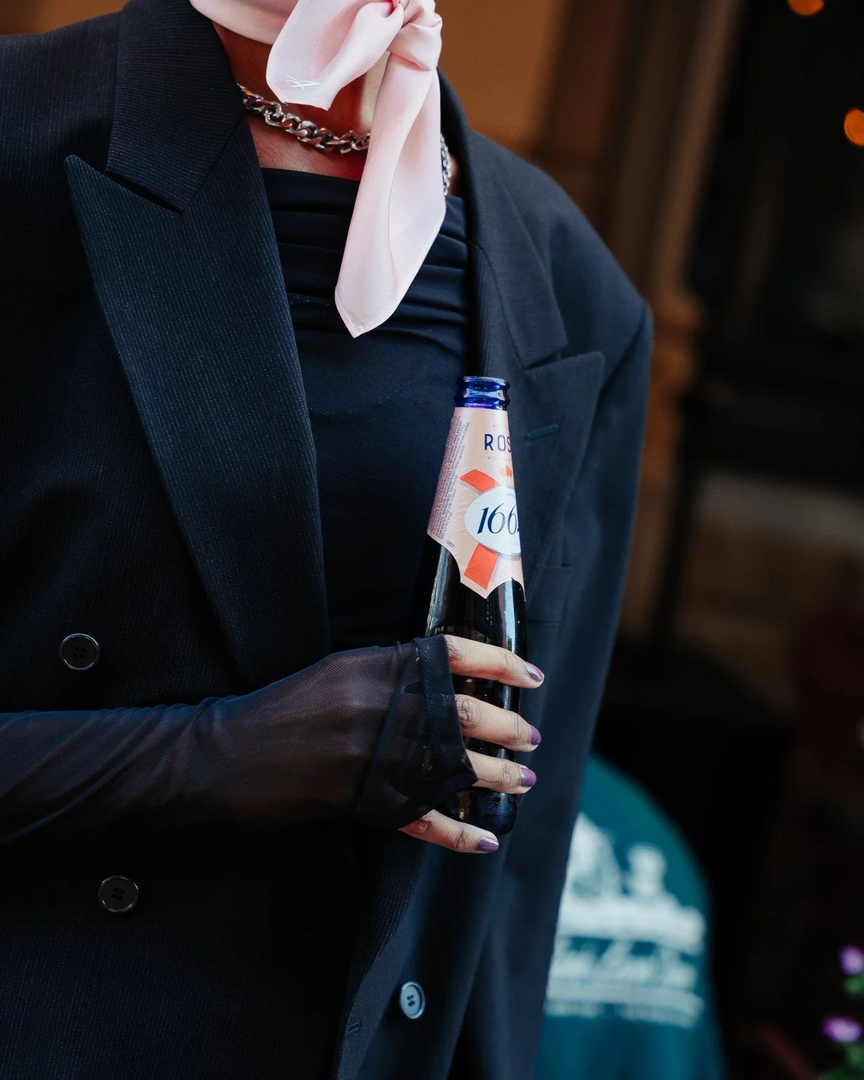 Person wearing a black blazer, black top, and sheer black glove holding a glass bottle of a beverage, with a white piece of cloth tied around their neck.
