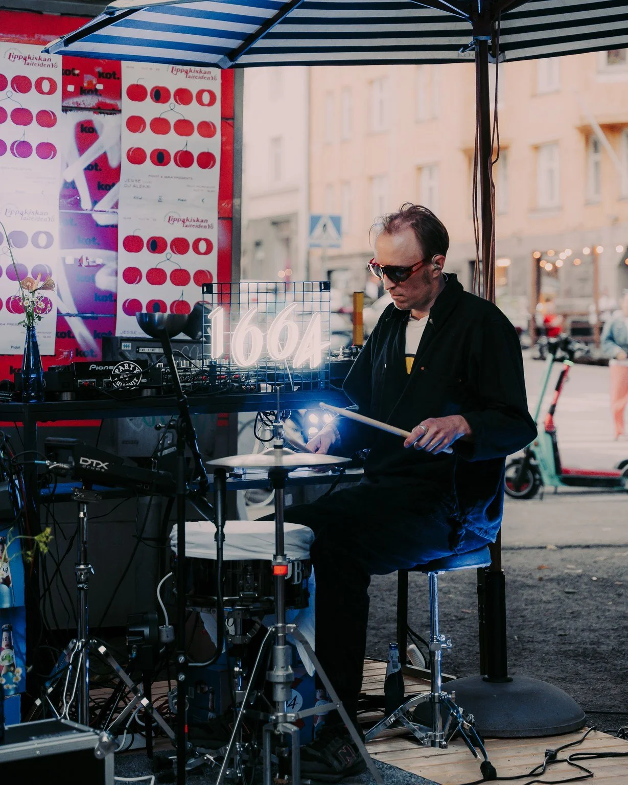 A musician performing at an outdoor event, seated at a drum set and surrounded by electronic equipment, with a neon sign displaying the number 1664. The background features posters and a street scene with buildings and electric scooters.