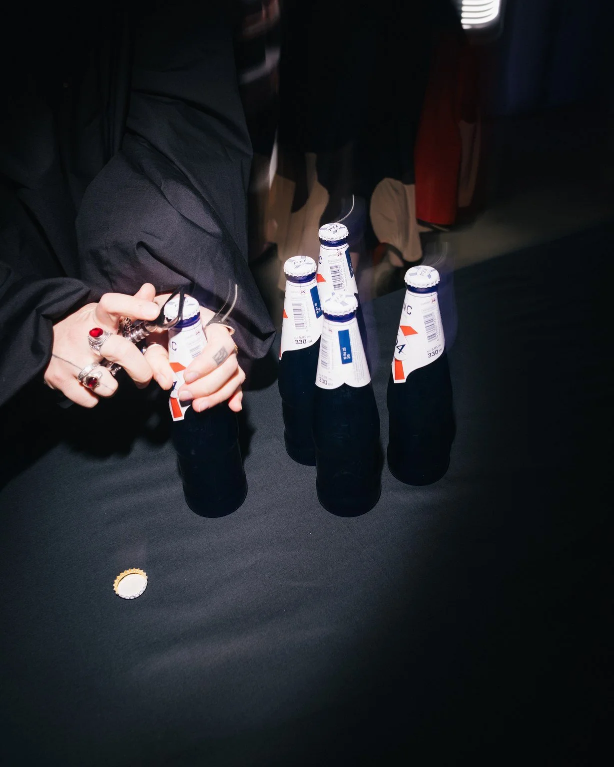 A person wearing multiple rings is holding a black bottle cap near a table with five bottles of beer arranged on it, with other people sitting in the background.