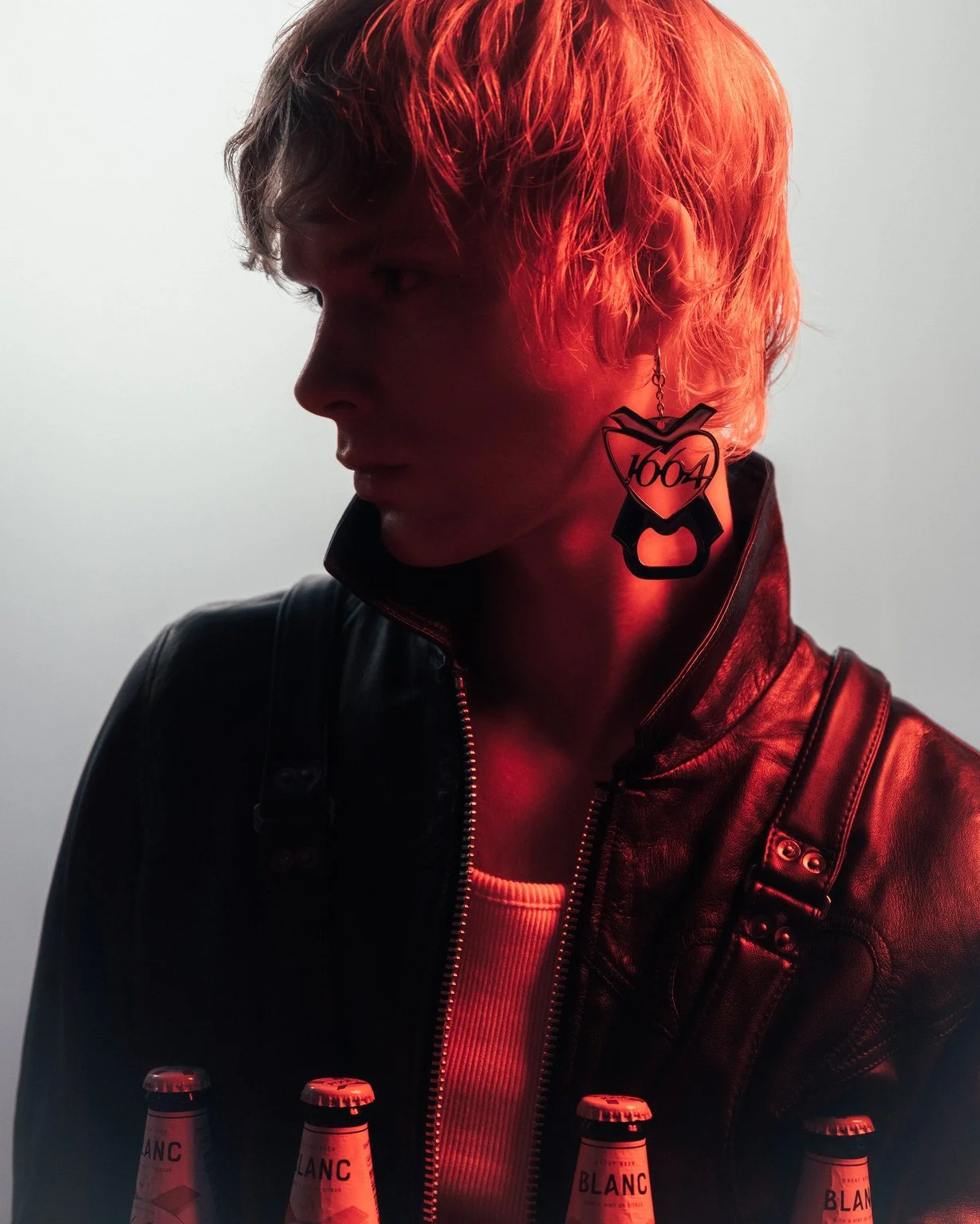 Portrait of a young person with wavy, orange hair wearing a black leather jacket and a white shirt, with large black earrings and standing in front of a plain background. The person has a side profile view with minimal lighting.