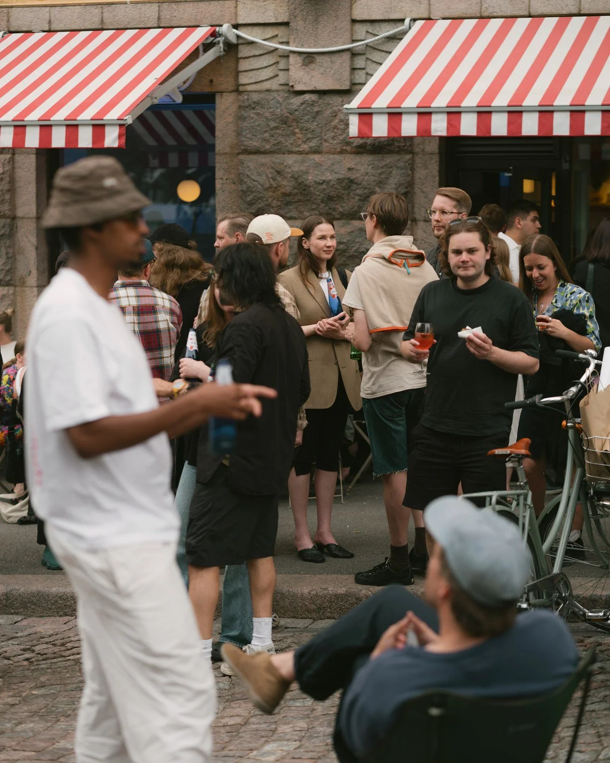 People gather outside a building with red and white striped awnings, socializing, drinking, and chatting. One person is sitting in a chair in the foreground, wearing a light-colored hat.