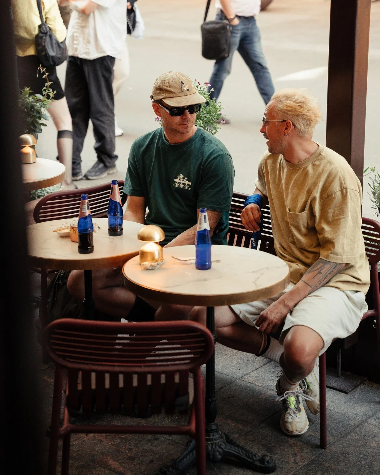 Two men are sitting at a table outside a restaurant, engaged in conversation. They each have three blue bottles and a small gold container on the table. The man on the left is wearing a green T-shirt, a beige cap, and sunglasses, while the man on the