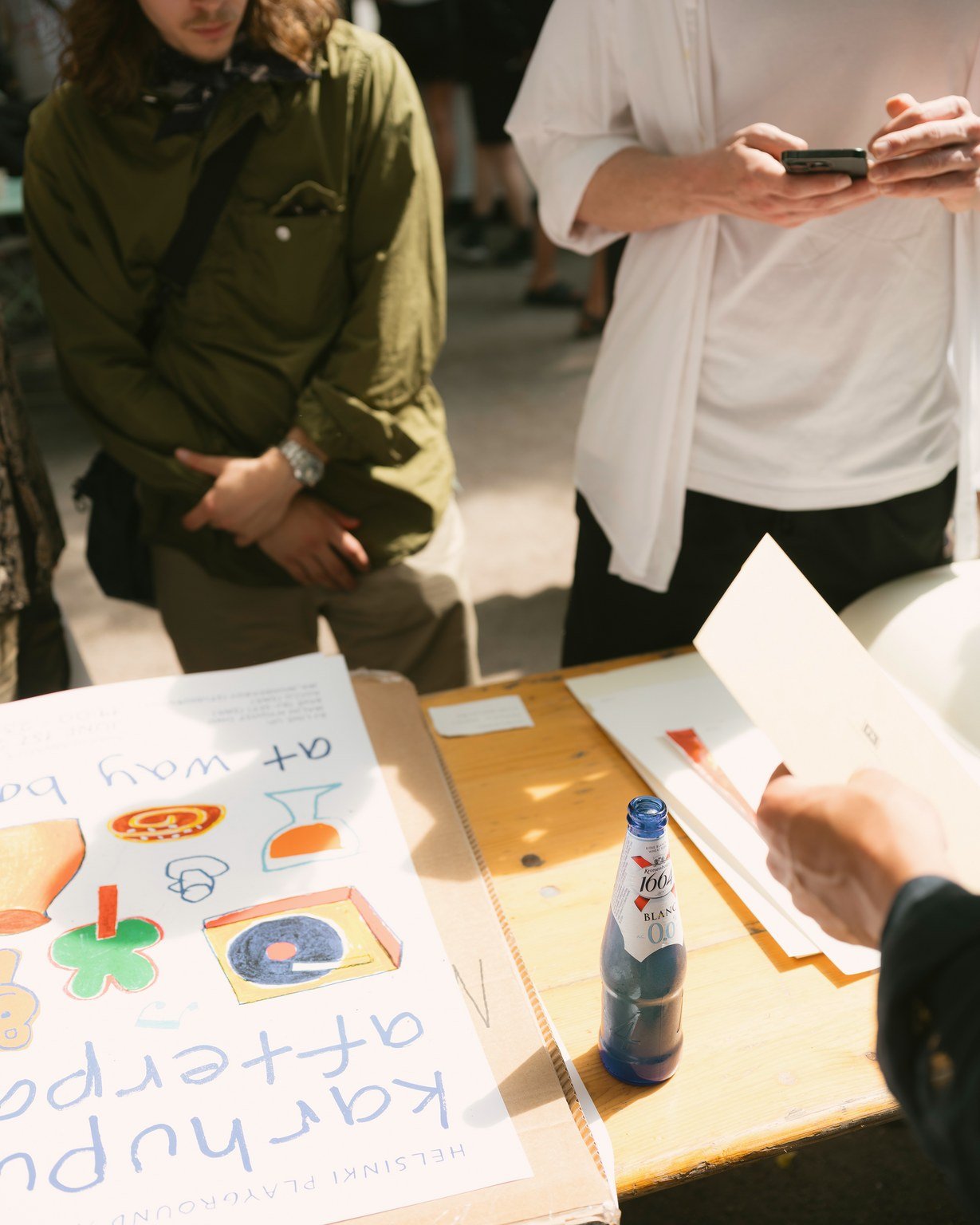 A tabletop scene showing a handmade poster with colorful drawings and text that reads 'KARASUPA + uplifting'; a person in black clothing holding a paper; a wooden table with a bottle of Asian sparkling water, and a notebook or pamphlet; two people st