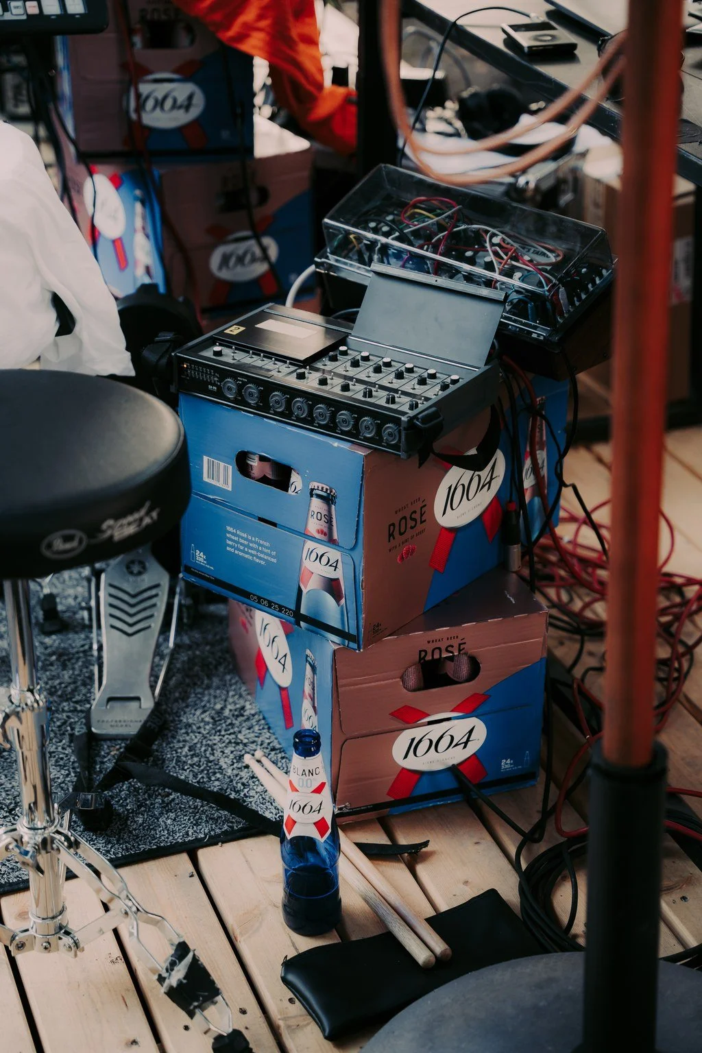 Music equipment setup on wooden floor including a drum pedal, a shaker, a bottle of beer, electronic music controller, and cables, with cardboard boxes and other gear in the background.