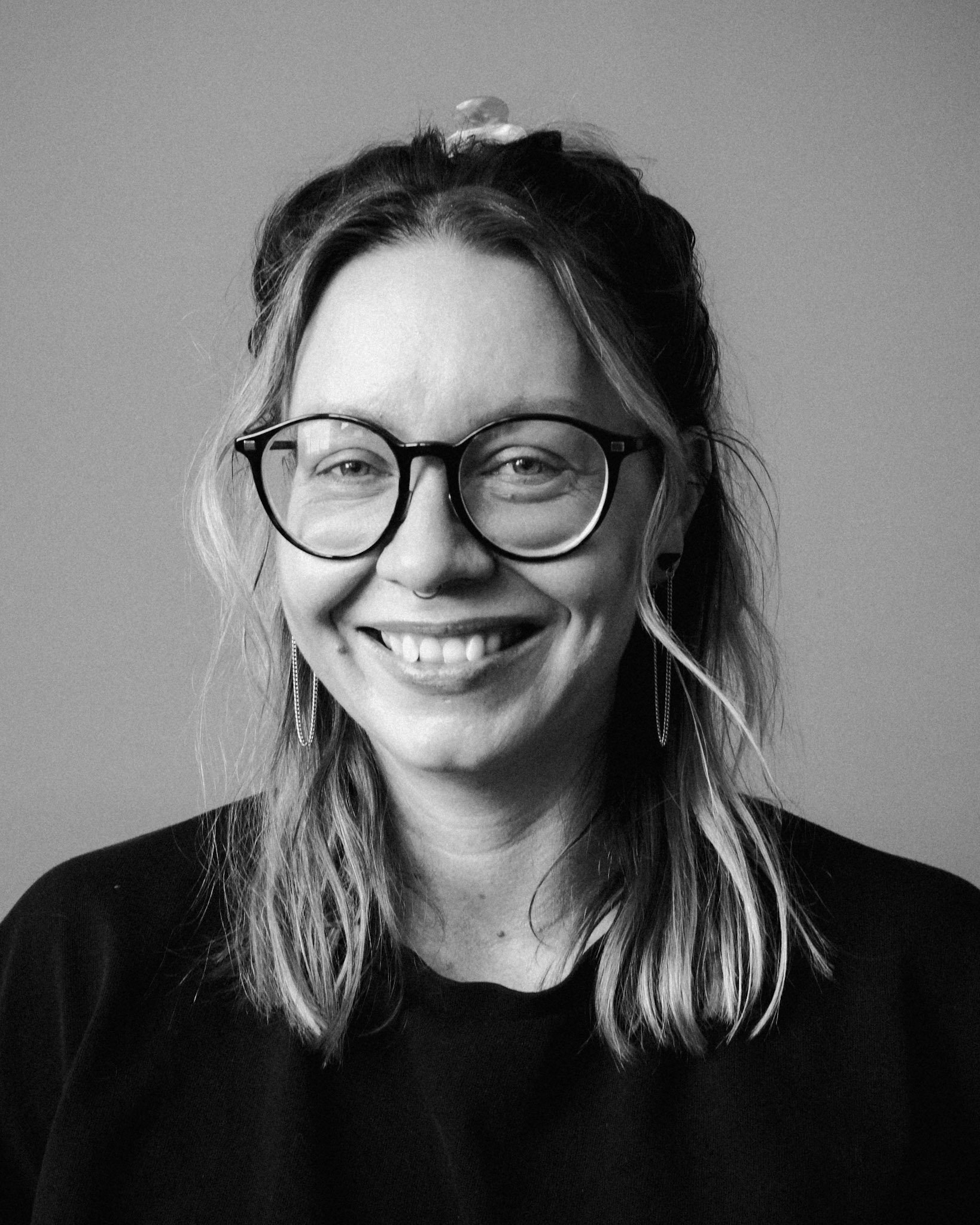 Black and white portrait of a woman smiling, wearing round glasses and a white collared shirt, with wavy hair and a nose piercing.