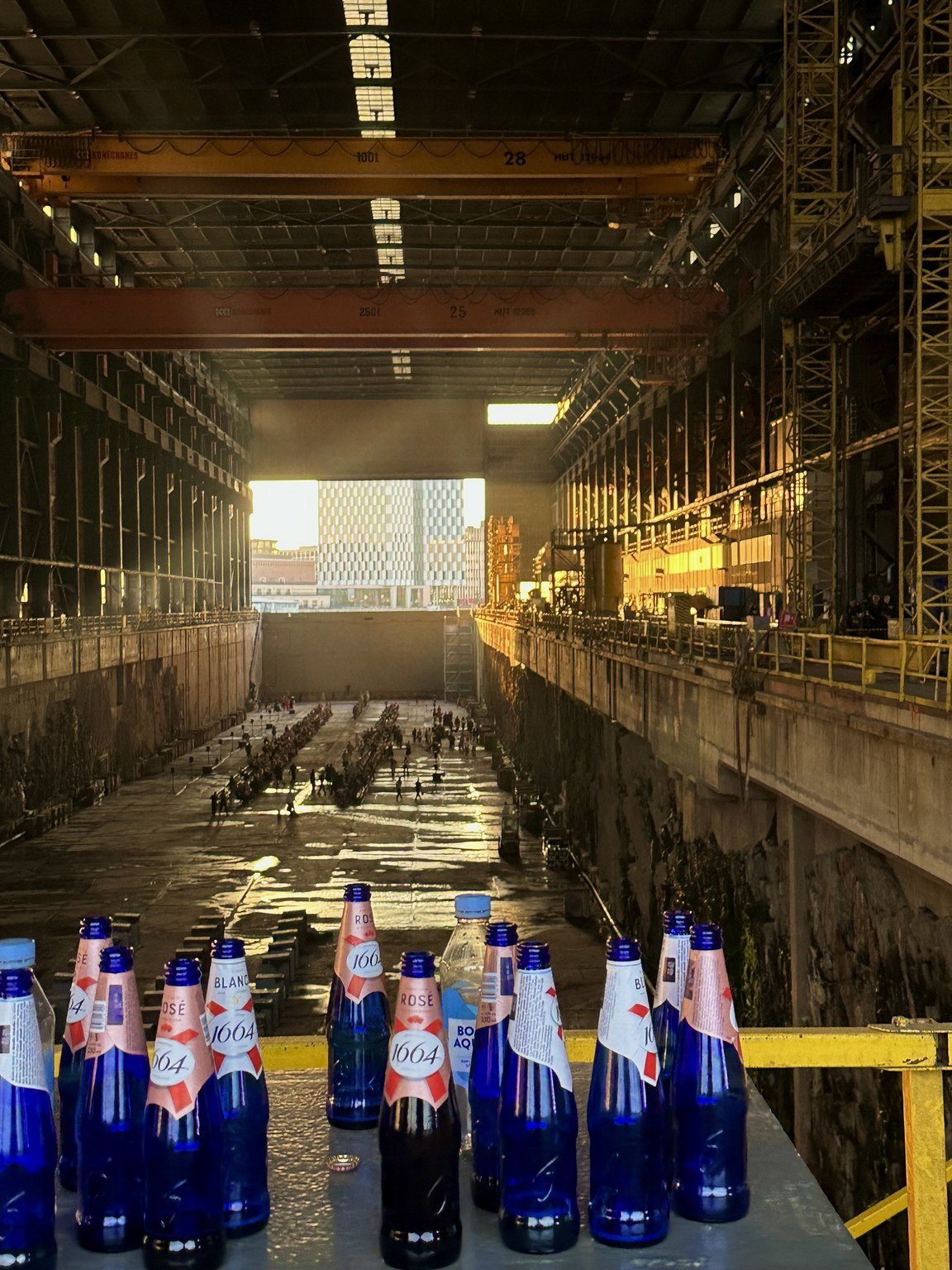 Blue glass bottles with pink and white labels are placed on a surface inside a large industrial building with scaffolding, machinery, and construction equipment, including a large overhead crane, with sunlight streaming through openings.