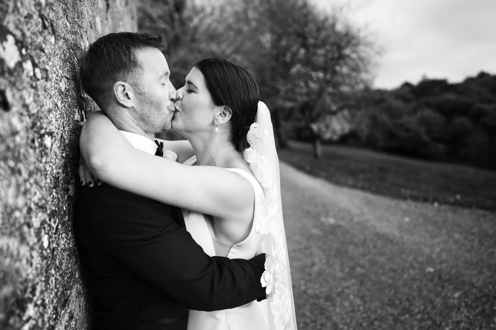 A black-and-white photo of a bride and groom kissing outdoors. The groom is dressed in a tuxedo, and the bride has her arms around his neck, wearing a sleeveless wedding dress with lace details and a veil. They are standing next to a textured wall in