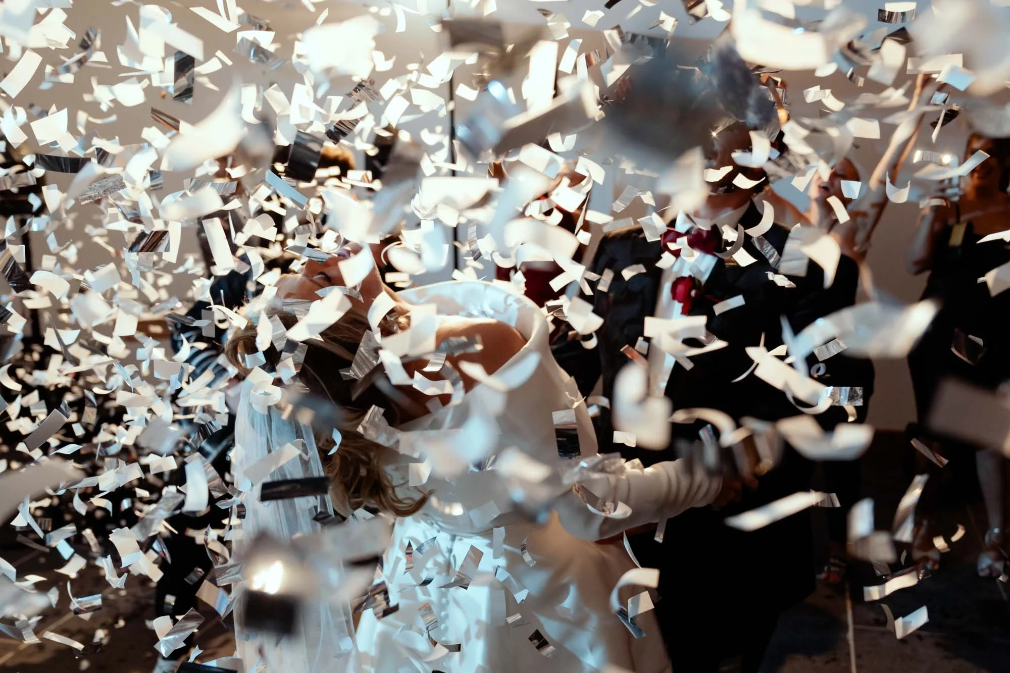 A wedding couple kissing as white confetti falls around them, with guests celebrating in the background.