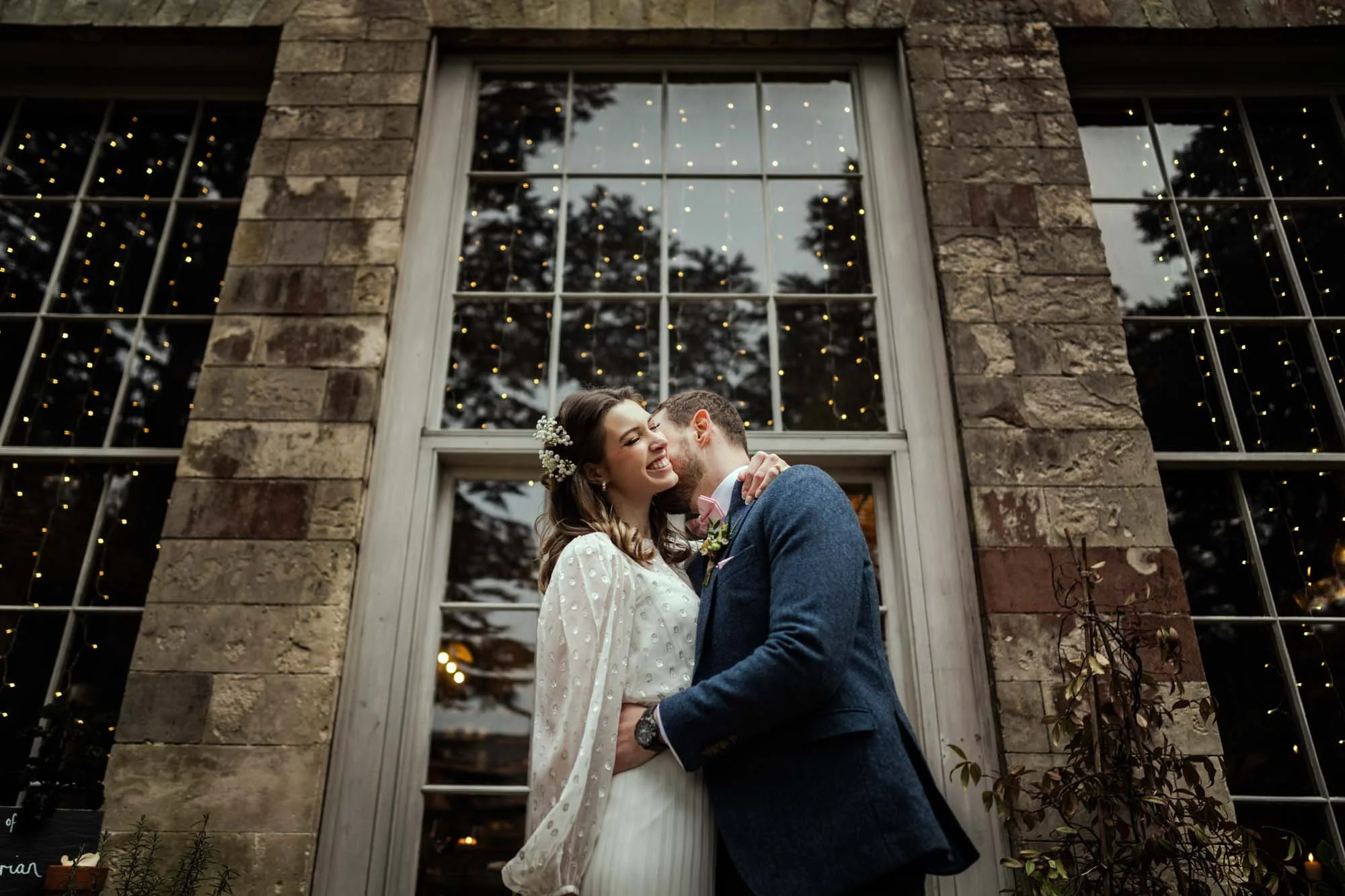 A happy couple sharing a kiss in front of a large window decorated with string lights and greenery, during a wedding celebration.
