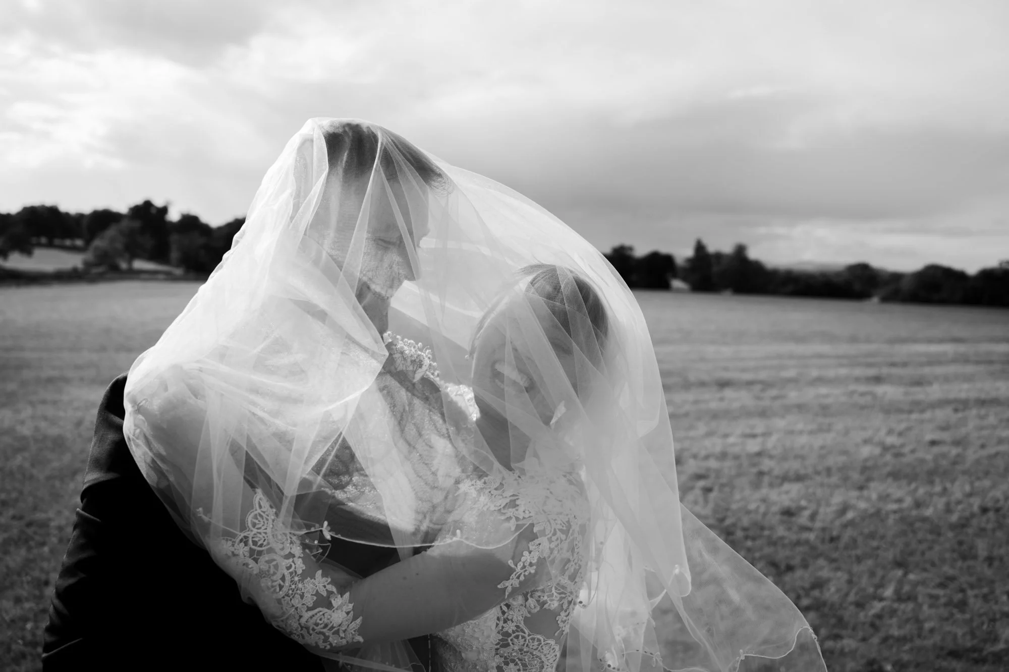 A bride and groom under a veil, embracing outdoors on a cloudy day with fields and trees in the background.