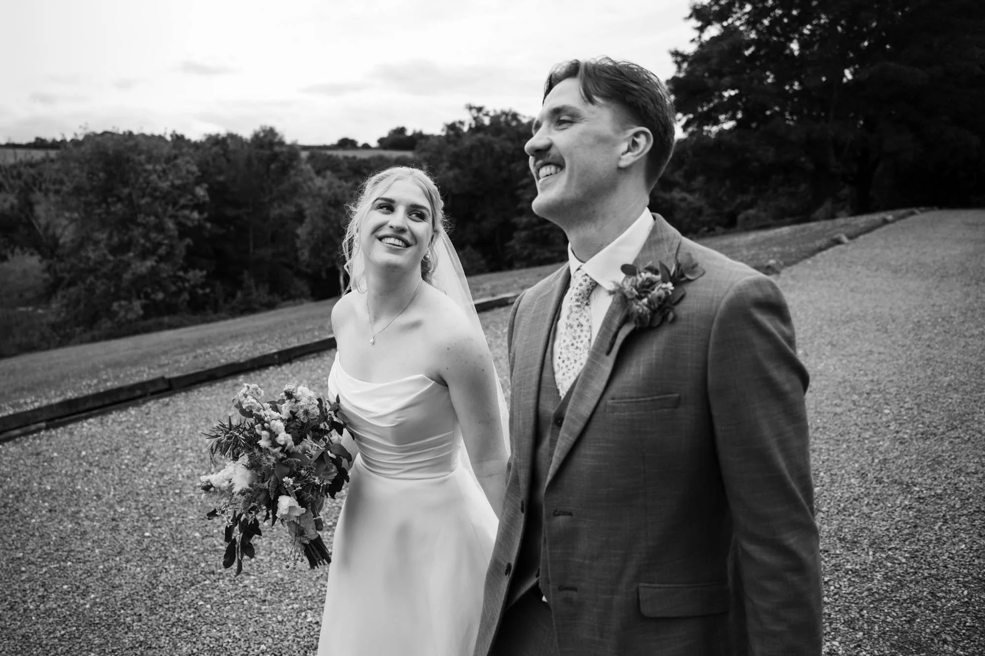 Black and white photo of a bride and groom outdoors, smiling and looking at each other, with trees in the background.