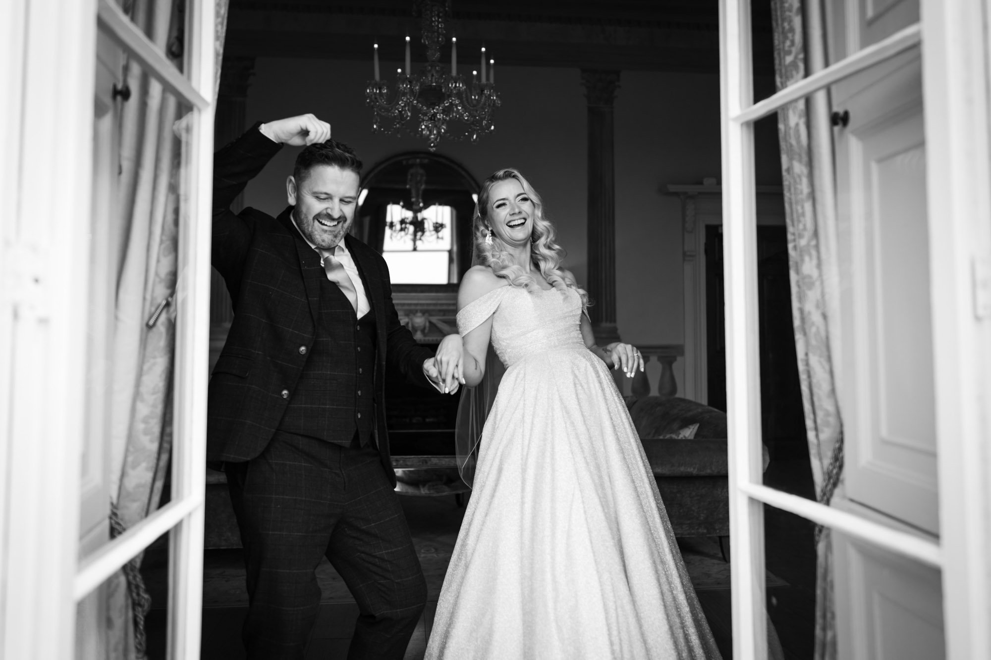 Black and white photo of a smiling bride in a wedding gown holding hands with a groom in a suit, dancing inside a decorated room visible through windowpanes.