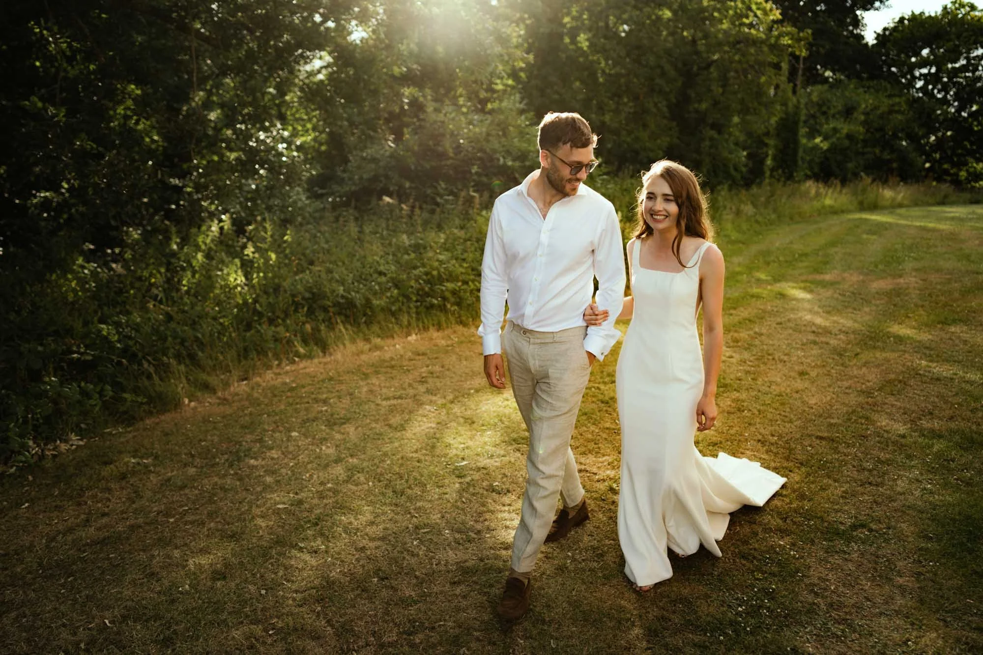 A smiling couple, with the man wearing a white shirt and beige pants, and the woman in a white dress, walking arm-in-arm on a grassy path in a park during sunset.