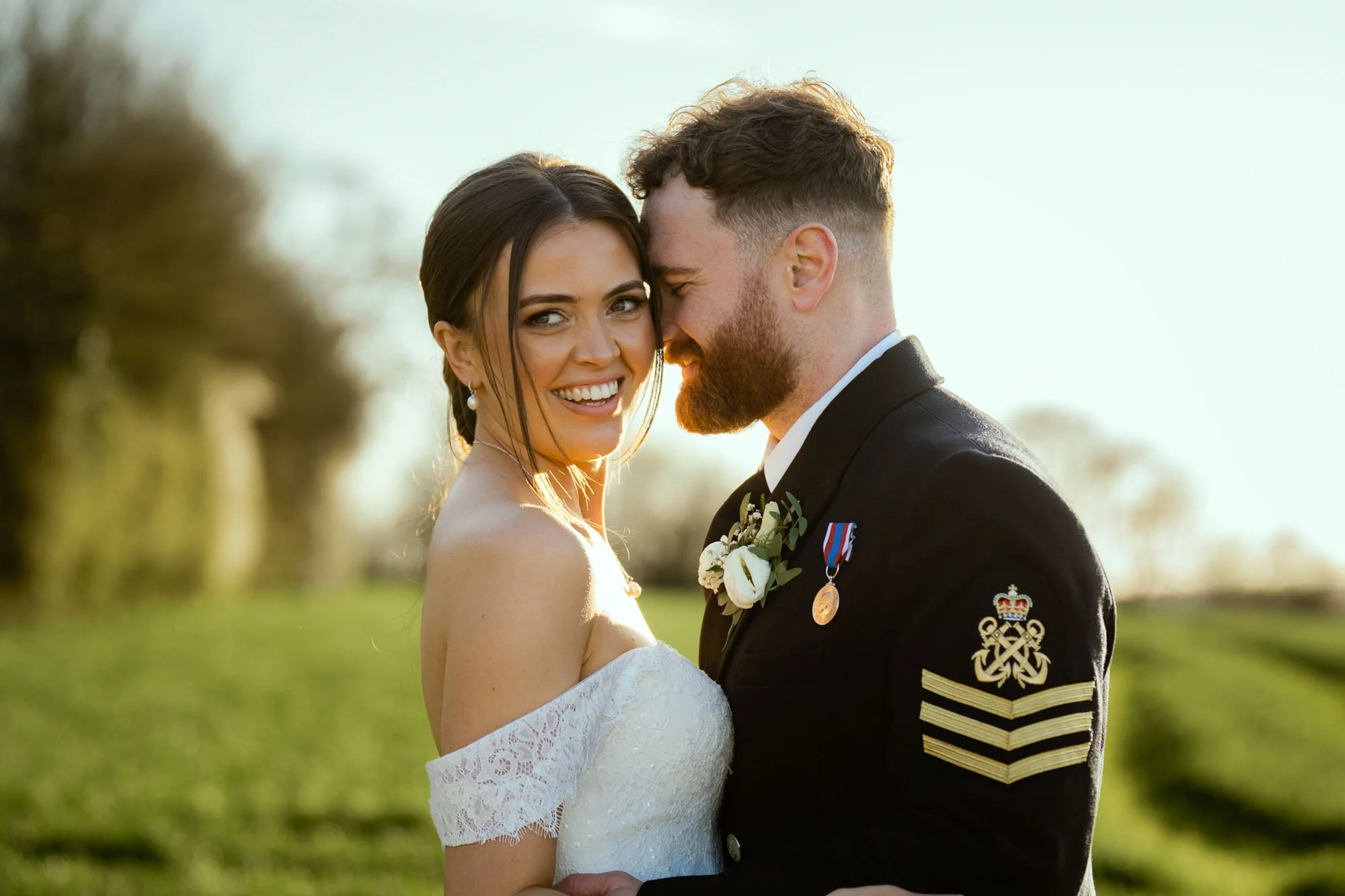 A newlywed couple smiling with their foreheads touching outdoors at sunset, the bride in an off-shoulder white wedding dress and the groom in a military uniform with medals and insignia.