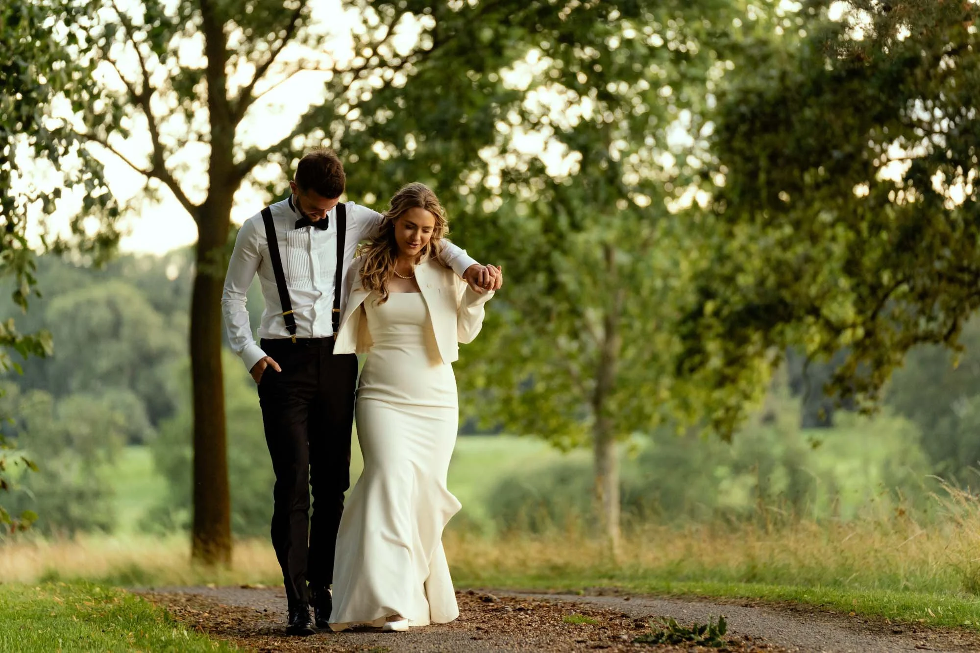 A bride and groom walking together outdoors on a dirt path surrounded by green trees, with the bride holding her dress as they enjoy a moment together.