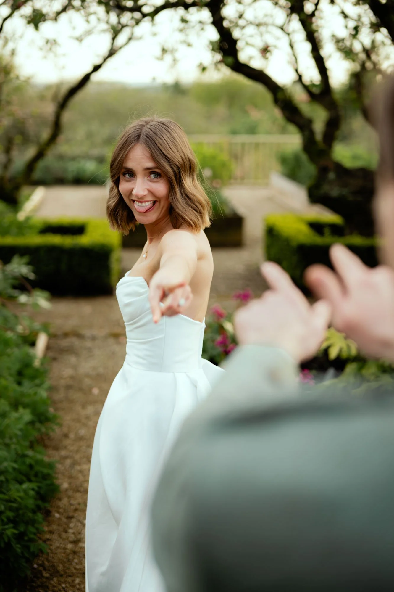 A woman in a white strapless dress smiling and pointing toward the camera in an outdoor garden setting.