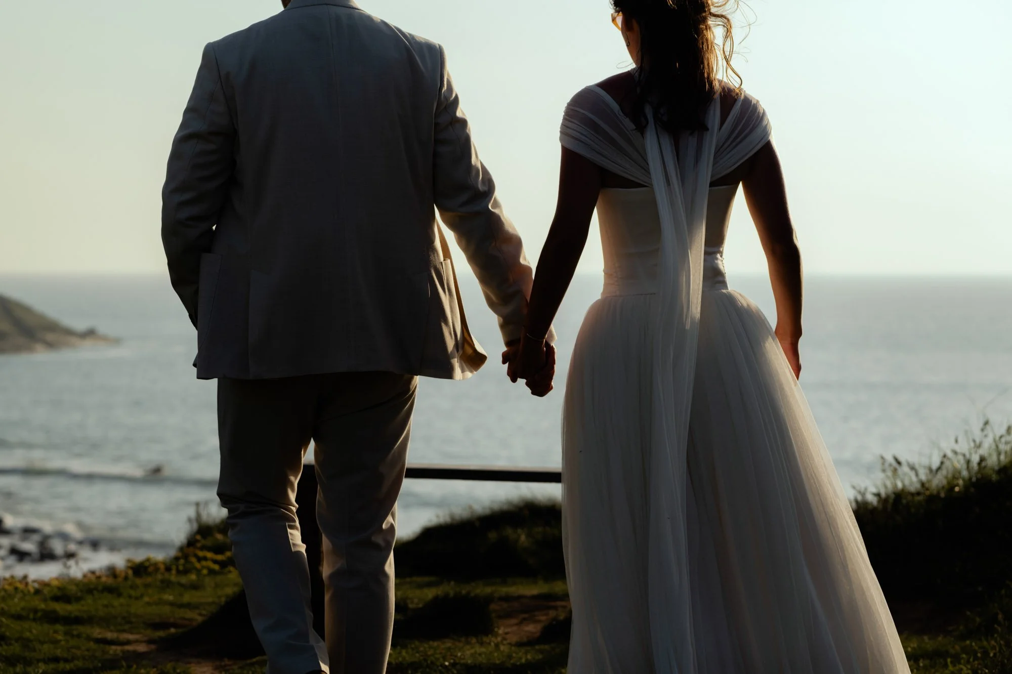 A couple holding hands, standing near the ocean at sunset, with the woman wearing a white wedding dress and the man in a gray suit.