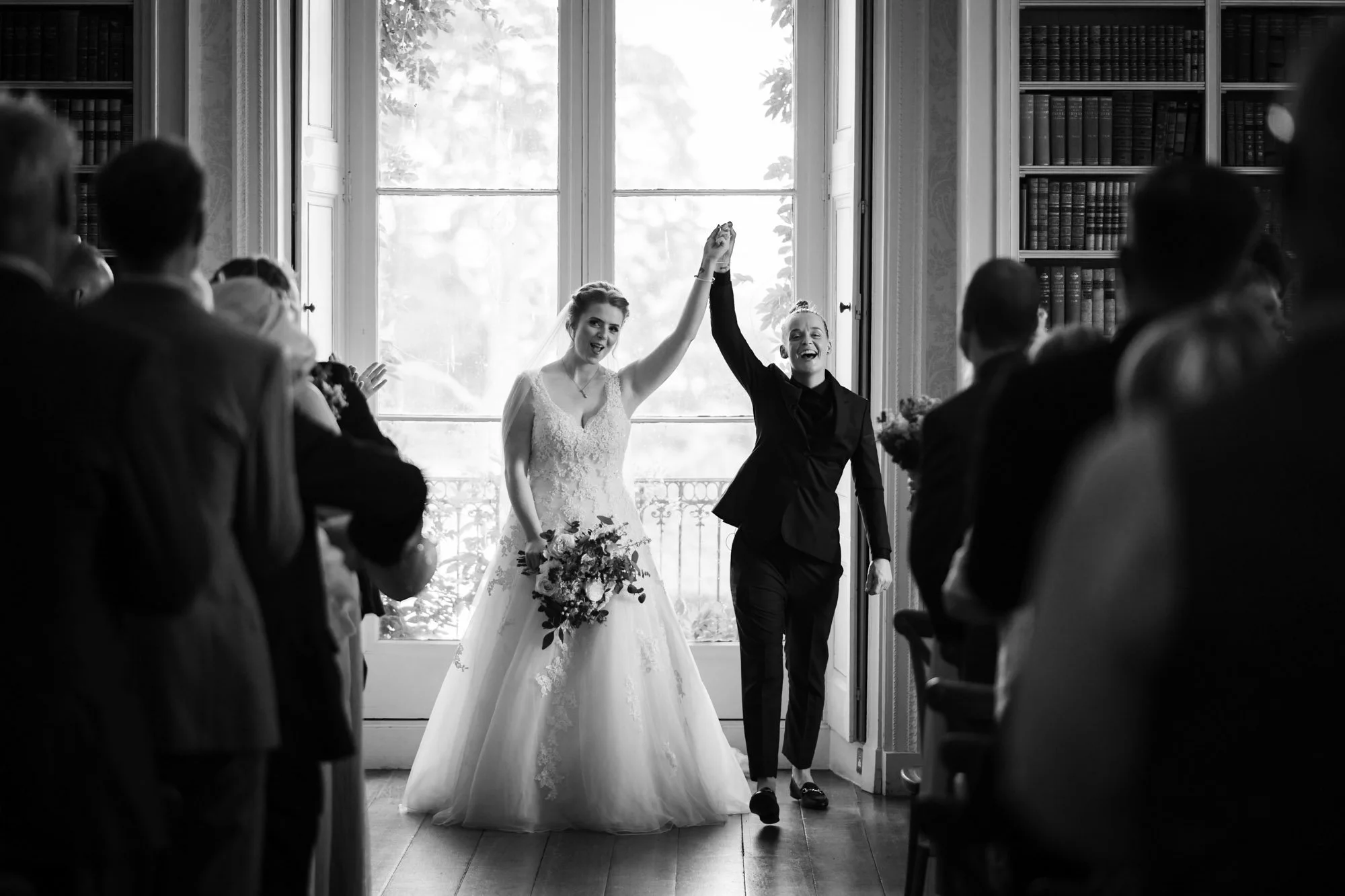 A black and white photo of two women, likely a bride and a wedding officiant, holding hands and smiling as they walk down the aisle in a room with a large window. The bride is in a wedding dress holding a bouquet, and both are surrounded by seated gu