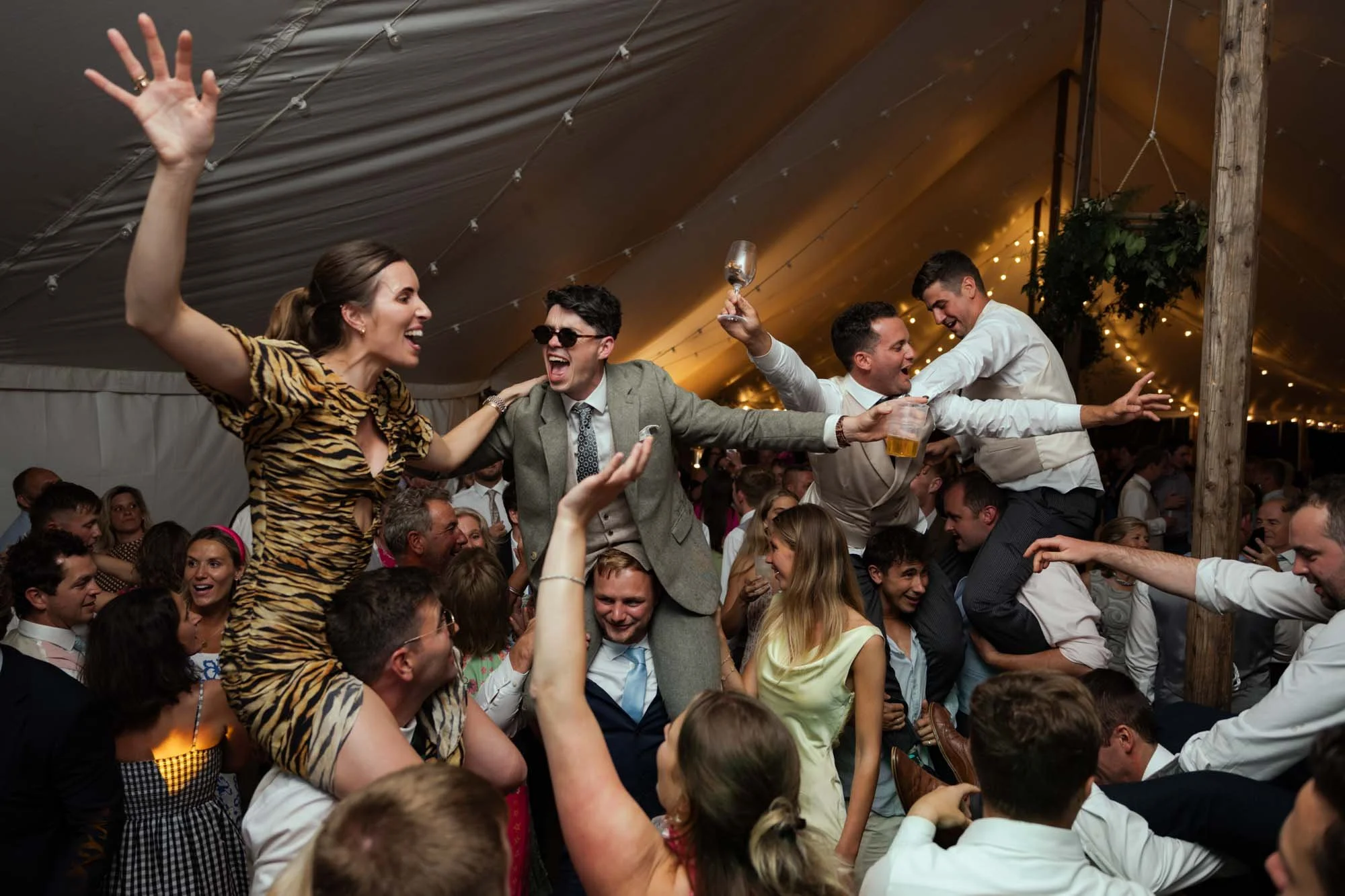 People celebrating at a lively event or party under a tent, with some being lifted in the air, holding drinks, and smiling.