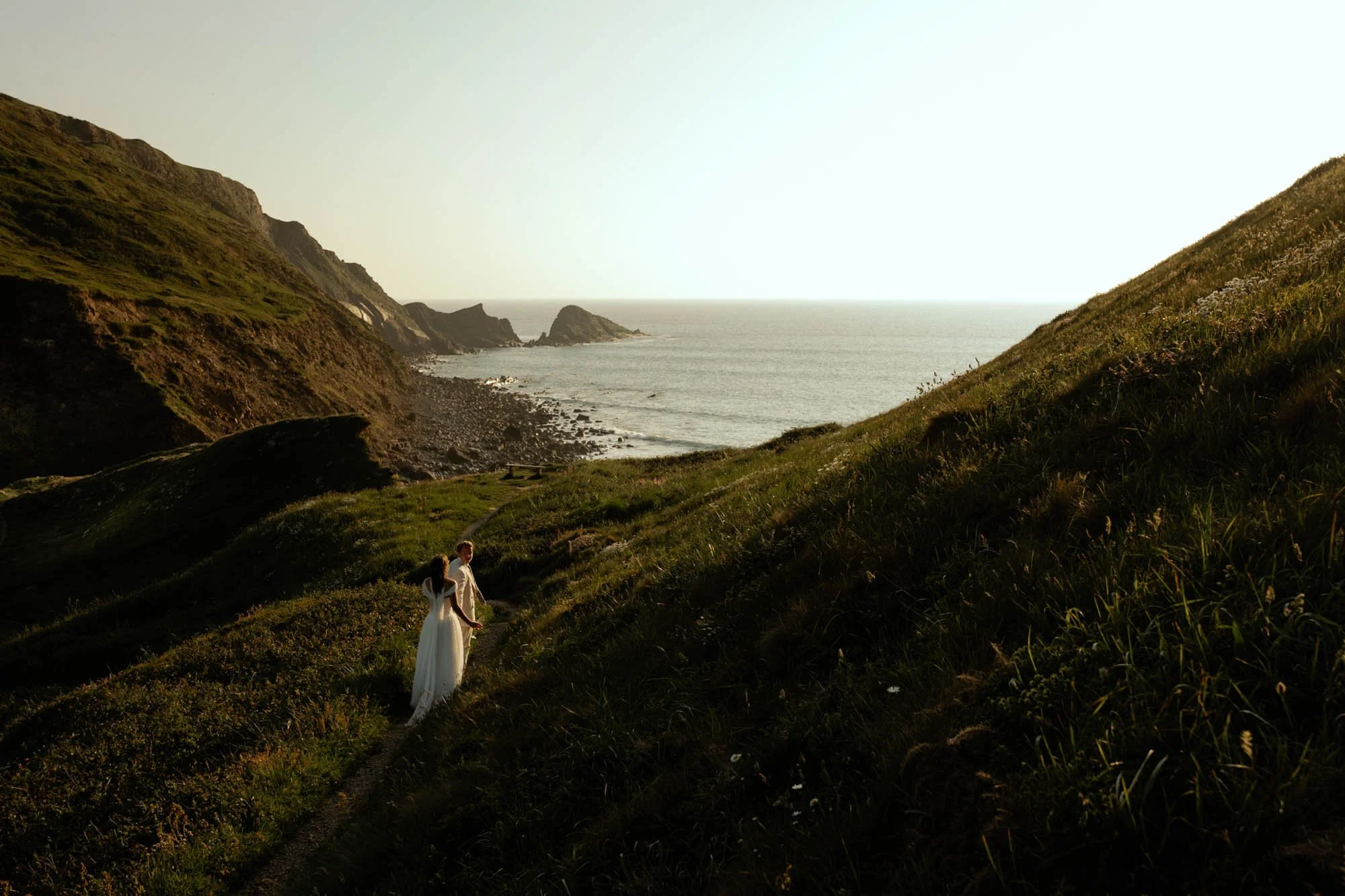 A couple holding hands walking along a grassy hillside overlooking the ocean during sunset.