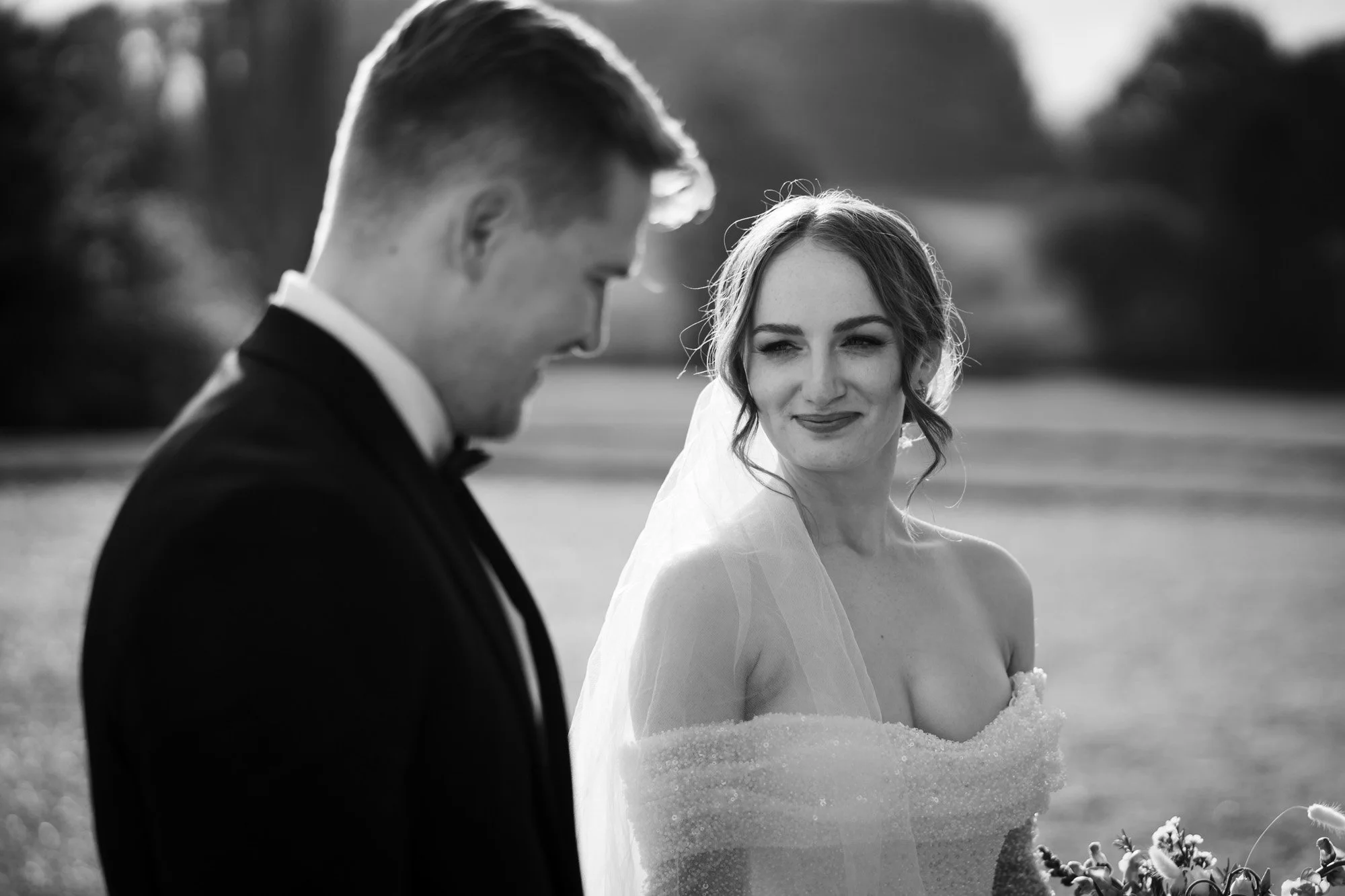 A black-and-white photograph of a bride and groom outdoors during their wedding. The groom, in a tuxedo with a bow tie, is on the left side, looking downward. The bride, on the right, is wearing a strapless wedding dress and veil, smiling softly as s