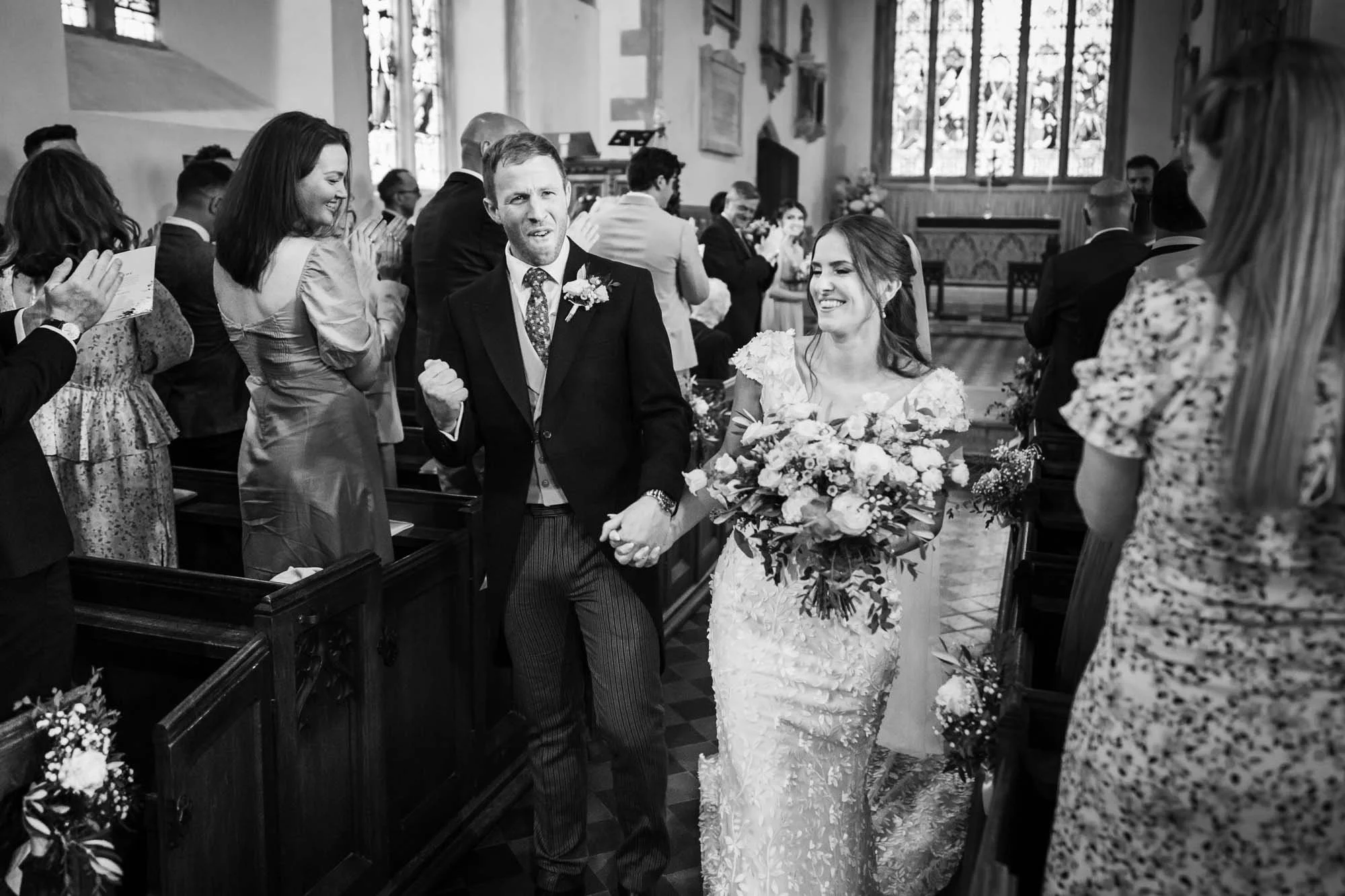 A bride and groom walking down the aisle holding hands, surrounded by seated and standing wedding guests inside a church.