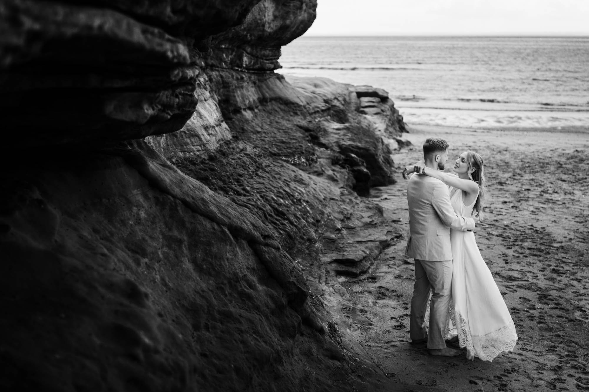 A couple dancing on the beach near rock formations, with the ocean in the background, during what appears to be a romantic moment.