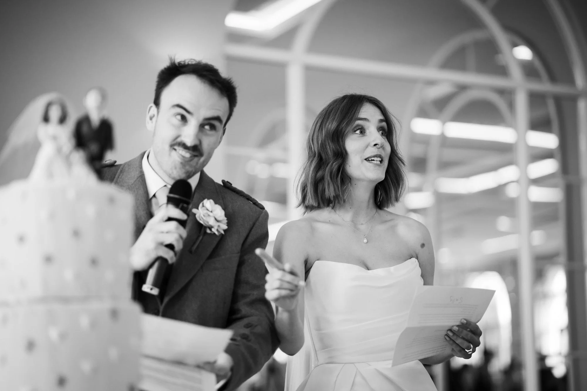 Black and white photo of a man and a woman at a wedding reception. The man is holding a microphone, smiling with one eye closed. The woman is holding a paper and looking surprised or emotional. The background features architectural windows.