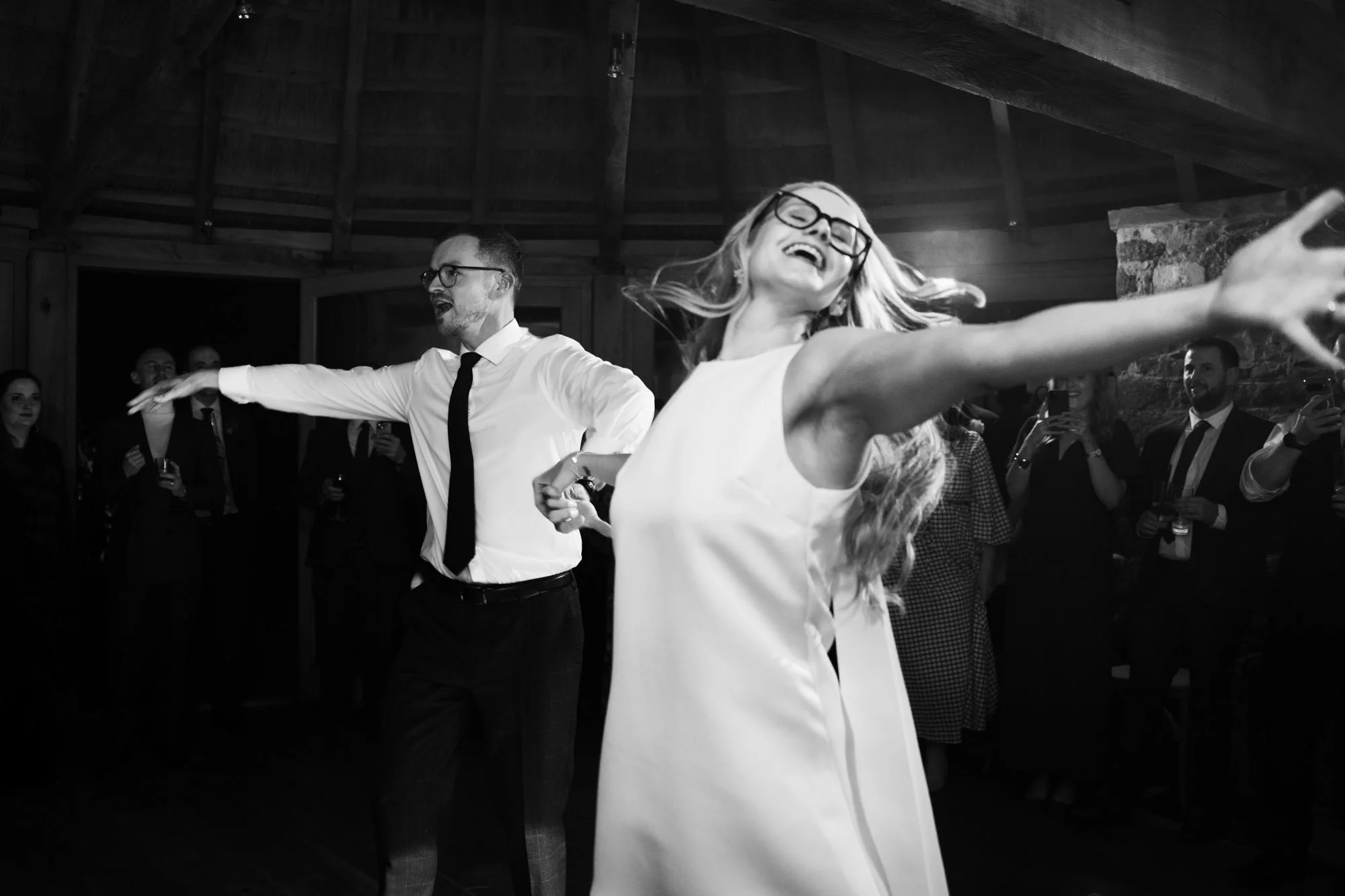 A woman in a sleeveless dress dancing and smiling with her arms outstretched, and a man in a white shirt and black tie dancing behind her, with guests watching and taking pictures in the background.