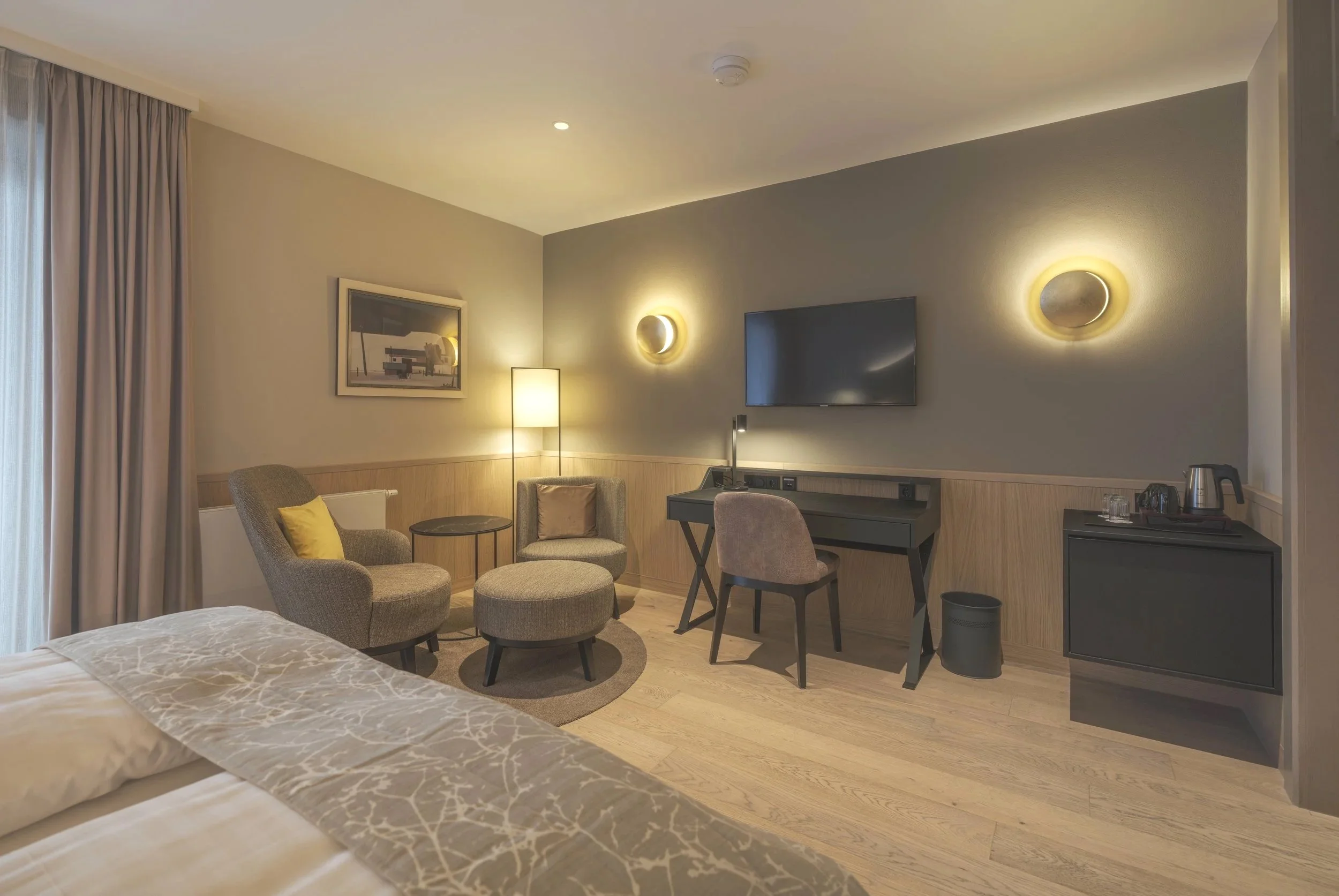 Hotelzimmer mit Doppelbett, Sitzbereich mit zwei Armchairs, einem kleinen Tisch, einem Schreibtisch mit Stuhl, Fernseher an der Wand, Stehlampe, Wanddekoration und einem Getränkeautomat