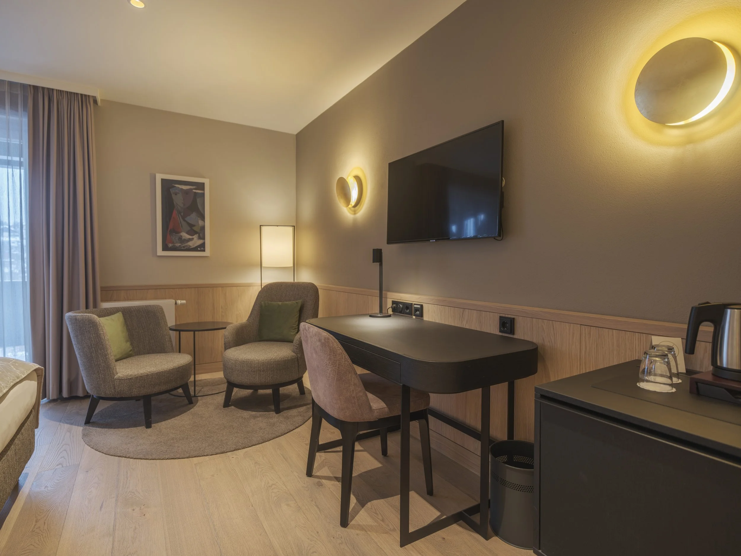 Gemütliches Hotelzimmer mit Sitzbereich, Fernseher, Stehlampe, Wall-Licht, Tisch, Stühlen, Teppich, Kaffeemaschine, Wasserkocher und Vorhängen.