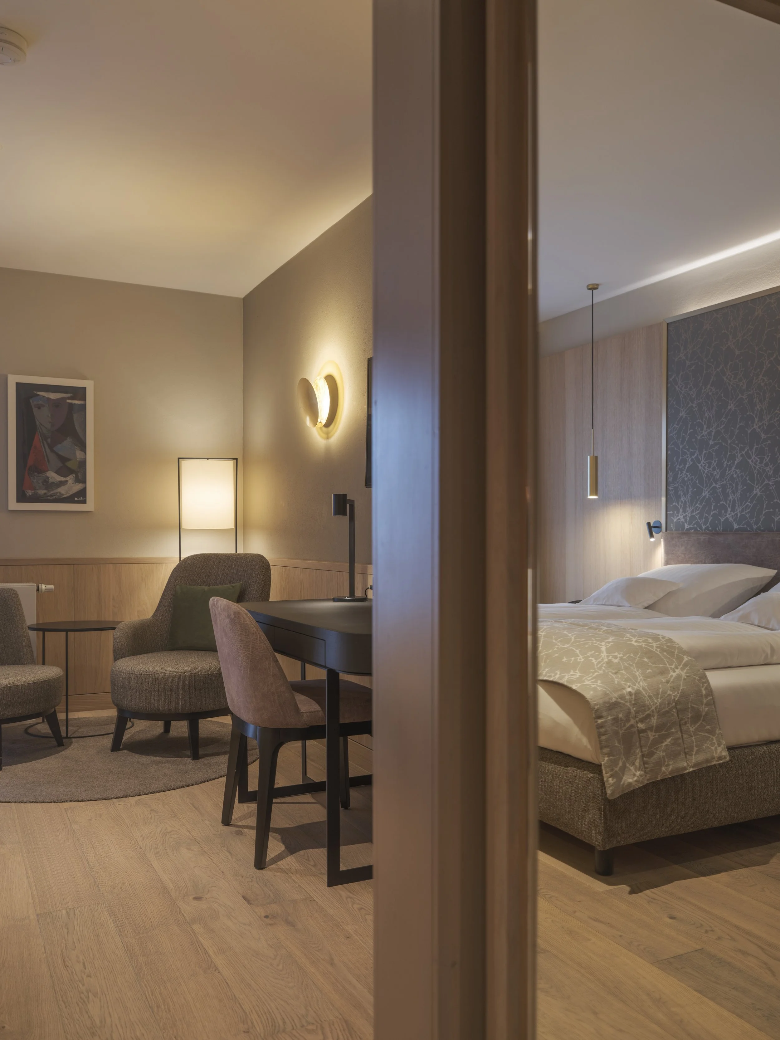 Zwei getrennte Hotelzimmer, die durch eine halb offene Trennwand voneinander getrennt sind. Auf der linken Seite befindet sich ein Wohnzimmer mit gemütlichen Sesseln, einem Tisch, einer Stehlampe, einem Wandbild und moderner Beleuchtung. Auf der rech