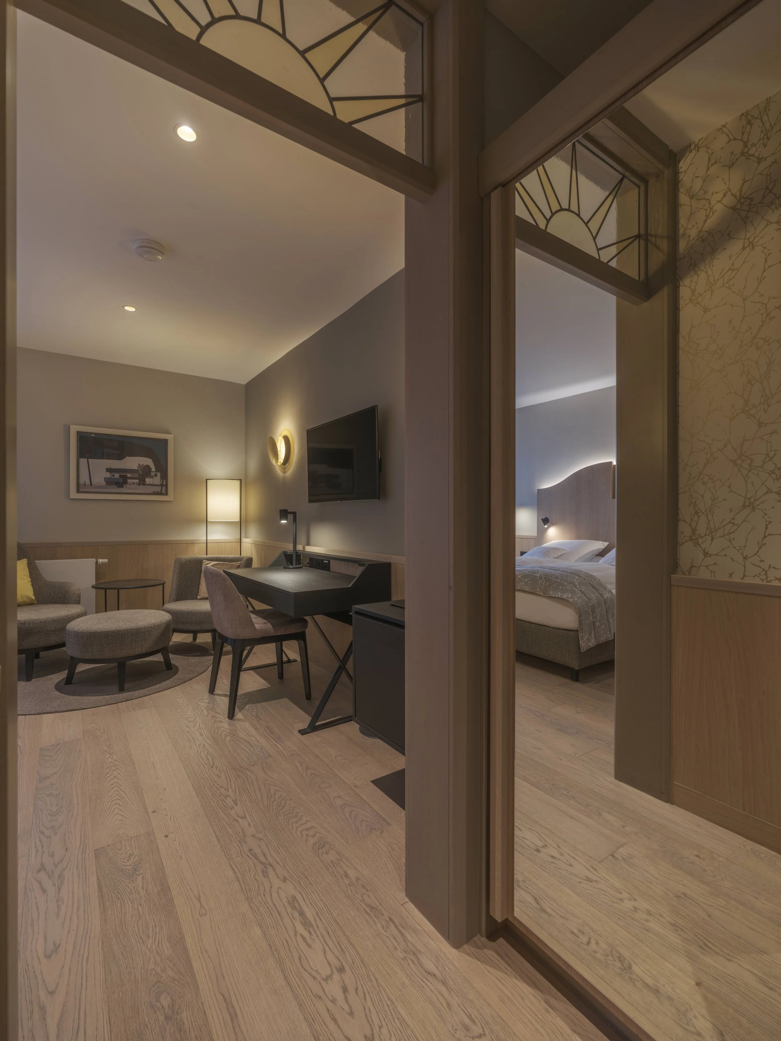 Innenaufnahme eines Hotels mit einem Schlafzimmer und einem Wohnzimmer, separaten Bereichen durch eine Tür, modern eingerichtet mit hellen Holzfußböden, einem Bett im Schlafzimmer und einem Sitzbereich im Wohnzimmer.