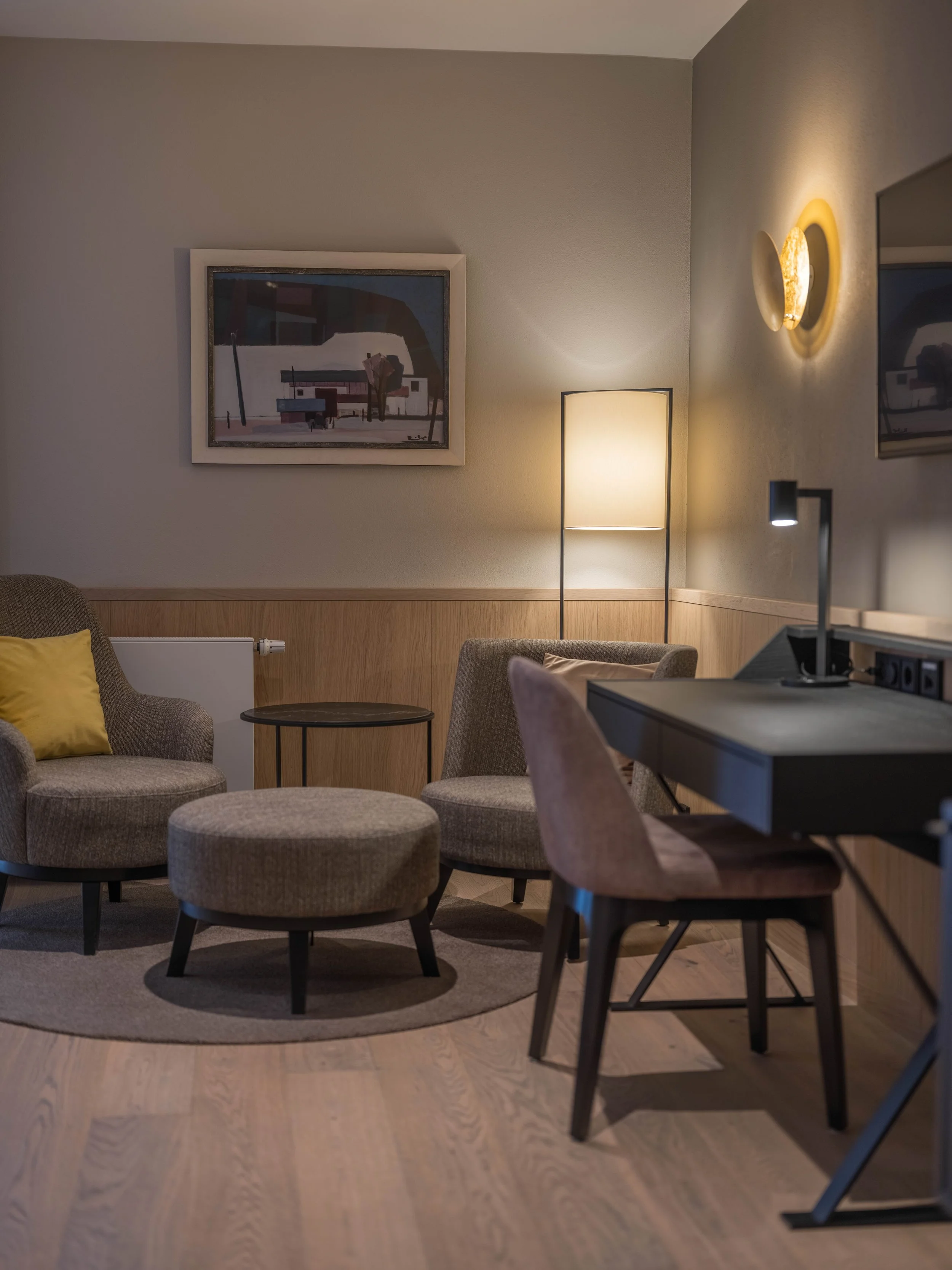 Gemütliches Wohnzimmer mit Stühlen, einem kleinen Tisch, Stehlampe, Wandlampen, Holzboden und Wanddekoration.