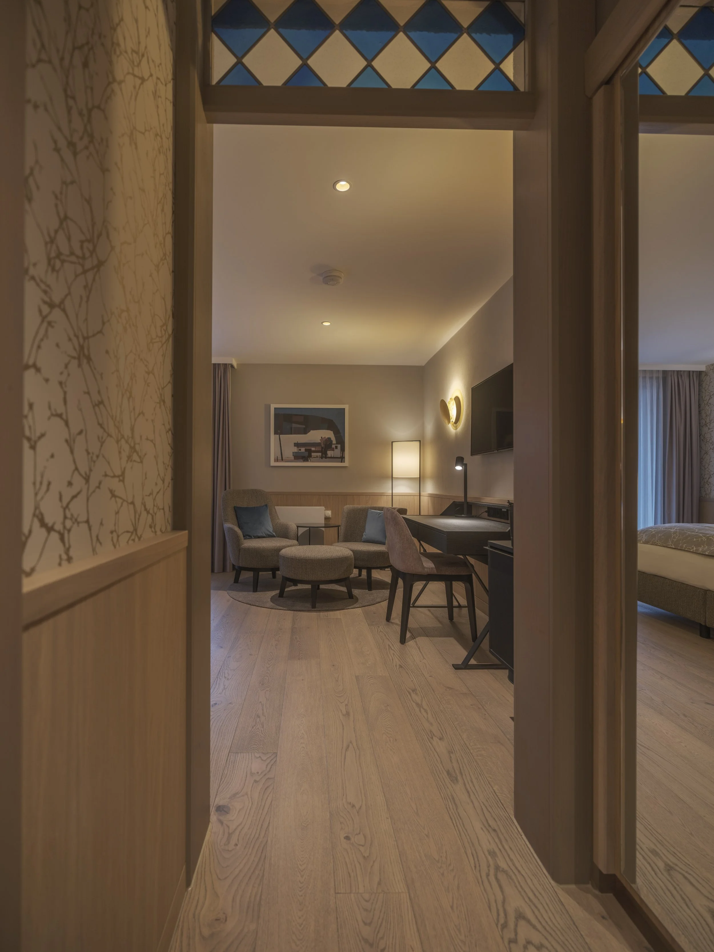 Ein Blick in ein modern eingerichtetes Hotelzimmer mit Sitzbereich, Schreibtisch, Fernseher und Schlafzimmer, warmes Licht, Holzboden, Außenglasfenster