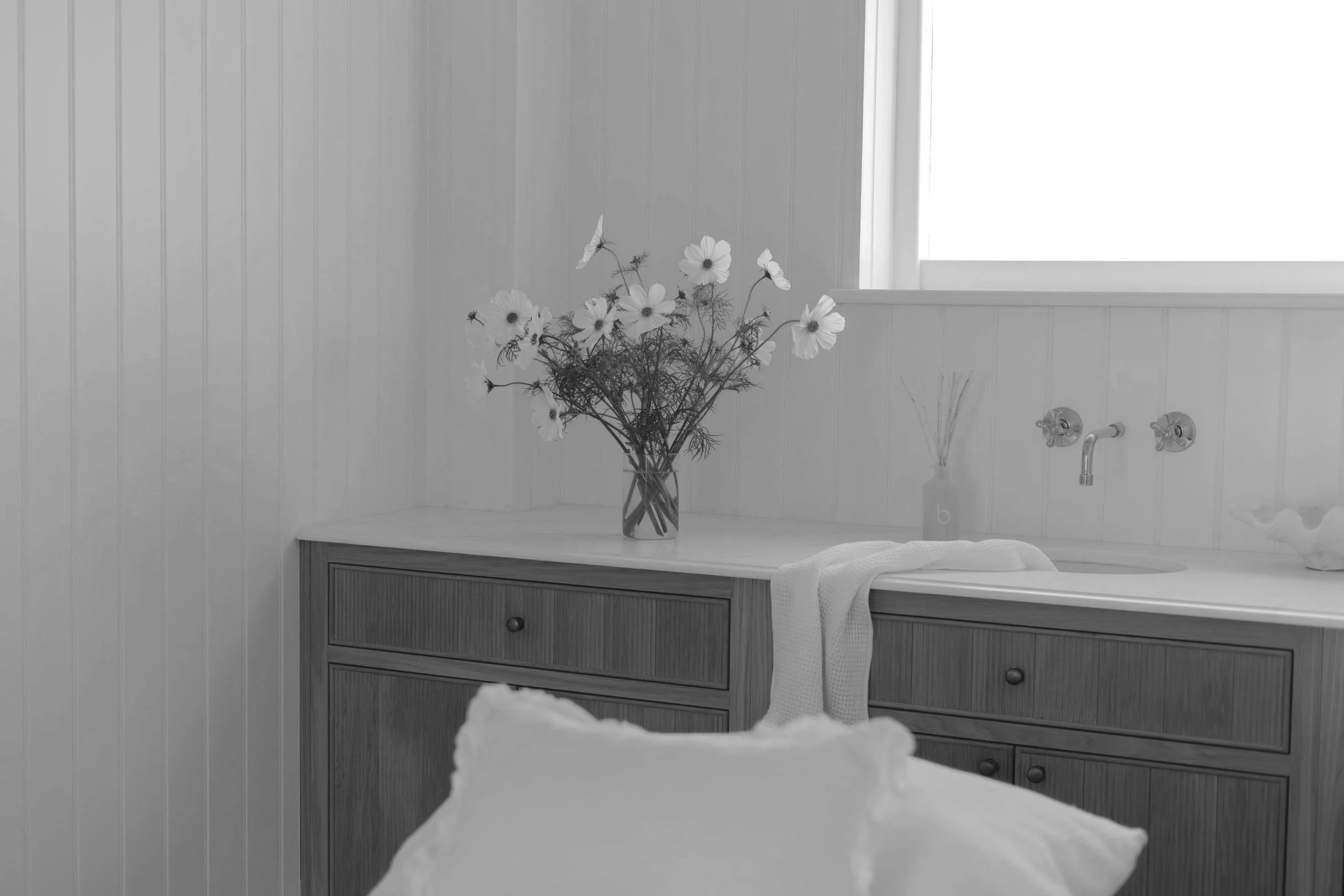 A bedroom with white paneled walls and a large window. On a wooden dresser, there is a glass jar filled with white flowers and greenery. A white towel is draped over the edge of the dresser, near the bottom right corner of the image.