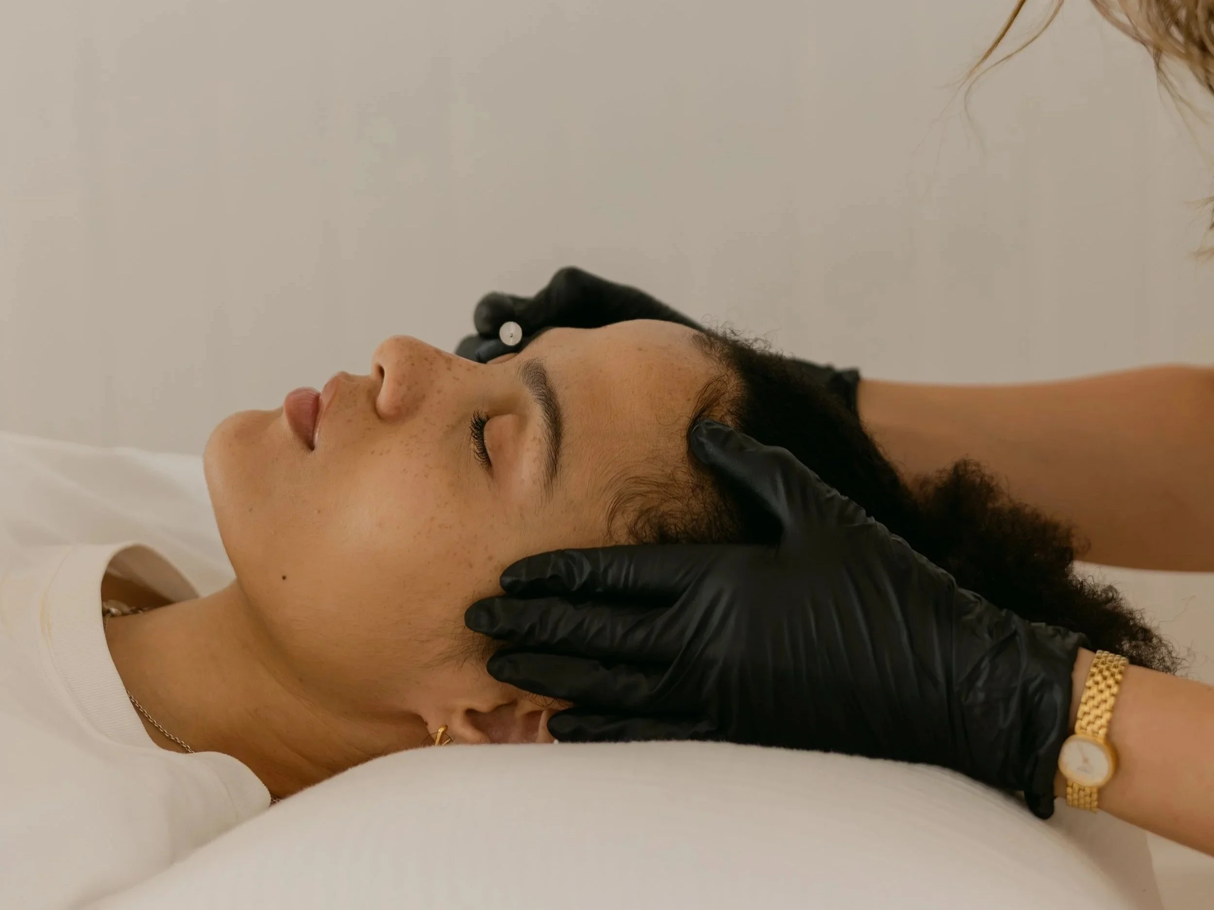 A woman lying down with eyes closed, receiving facial treatment from a skincare professional wearing black gloves.