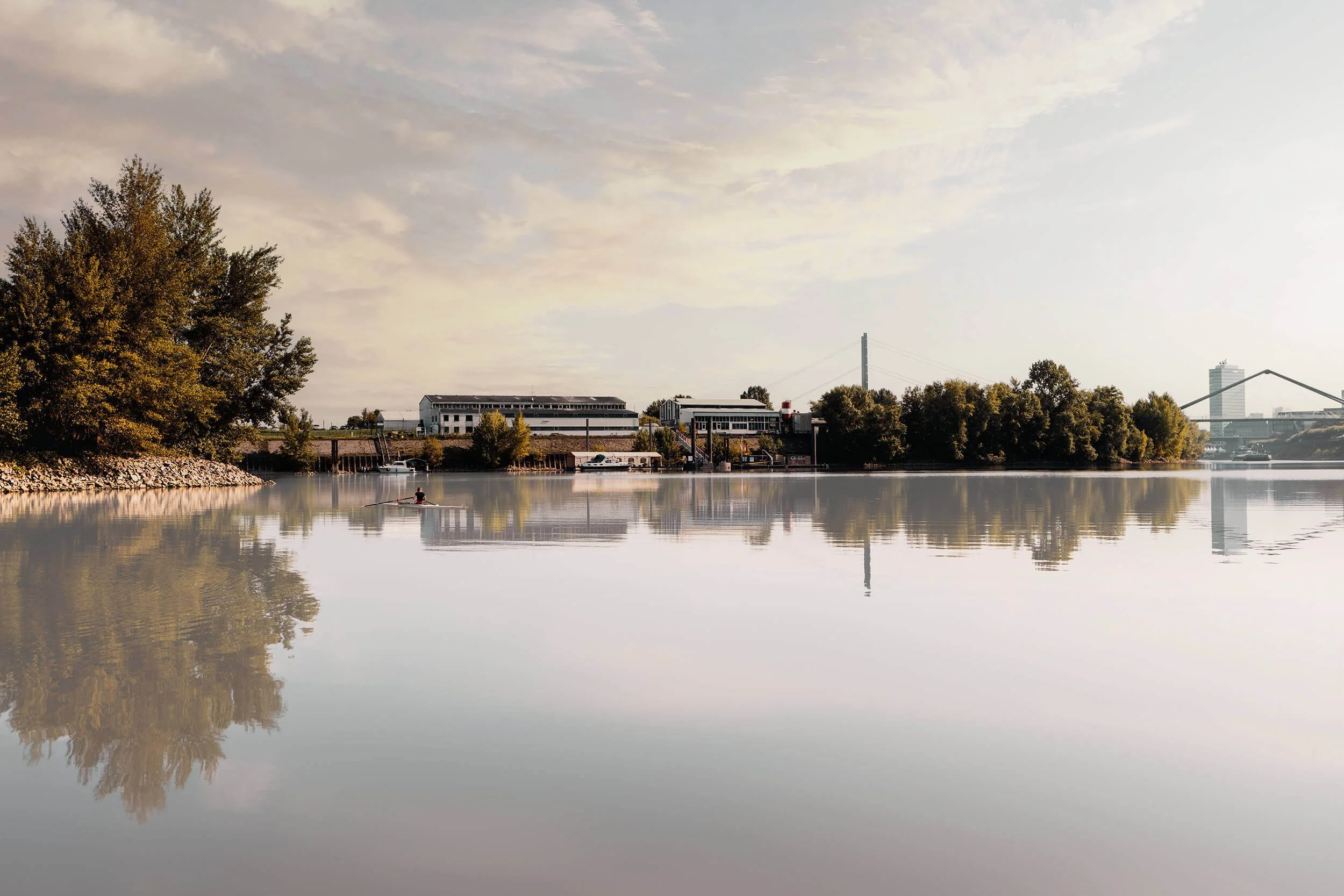 Romantischer Blick auf die Werkhallen der Kunstgießerei Rolf Kayser im Düsseldorfer Hafen. Im Hintergrund die Rheinkniebrücke sowie die Brücke am Medienhafen und das Mannesmann-Hochhaus. Auf dem Wasser eine Ruderin des Ruderclubs Germania.