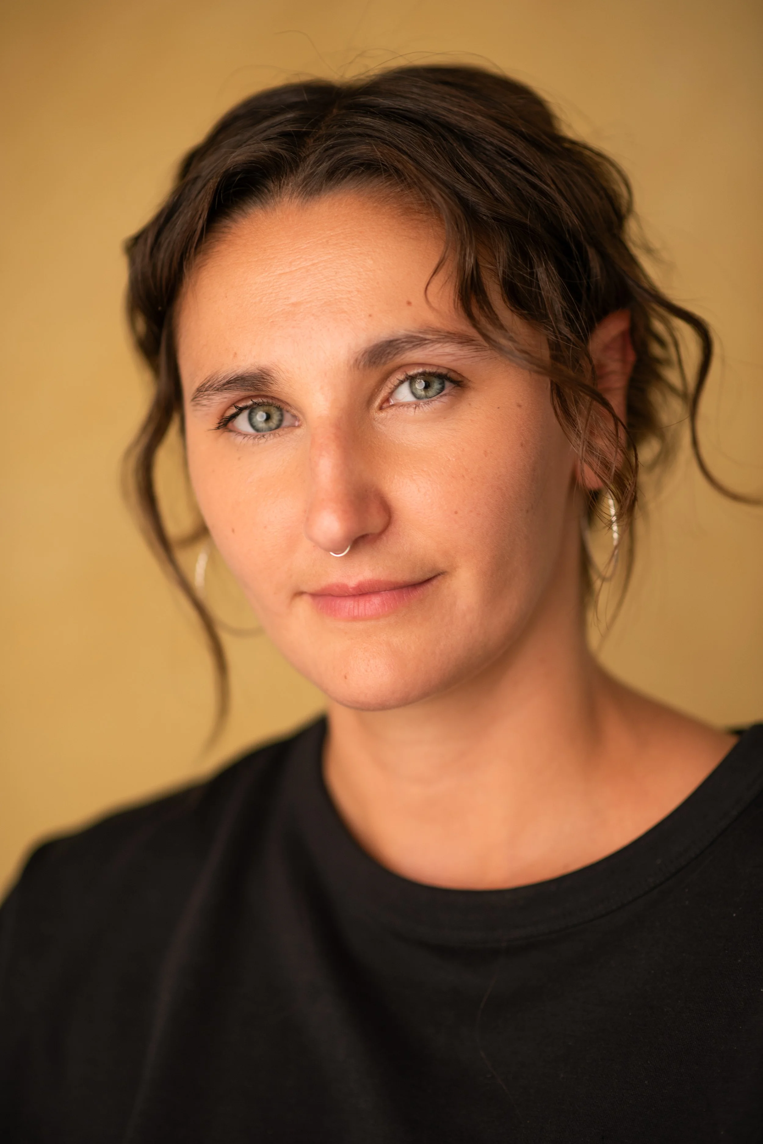 Close-up portrait of a woman with short, wavy brown hair, light skin, blue eyes, a septum piercing, and wearing a black top, against a yellow background.