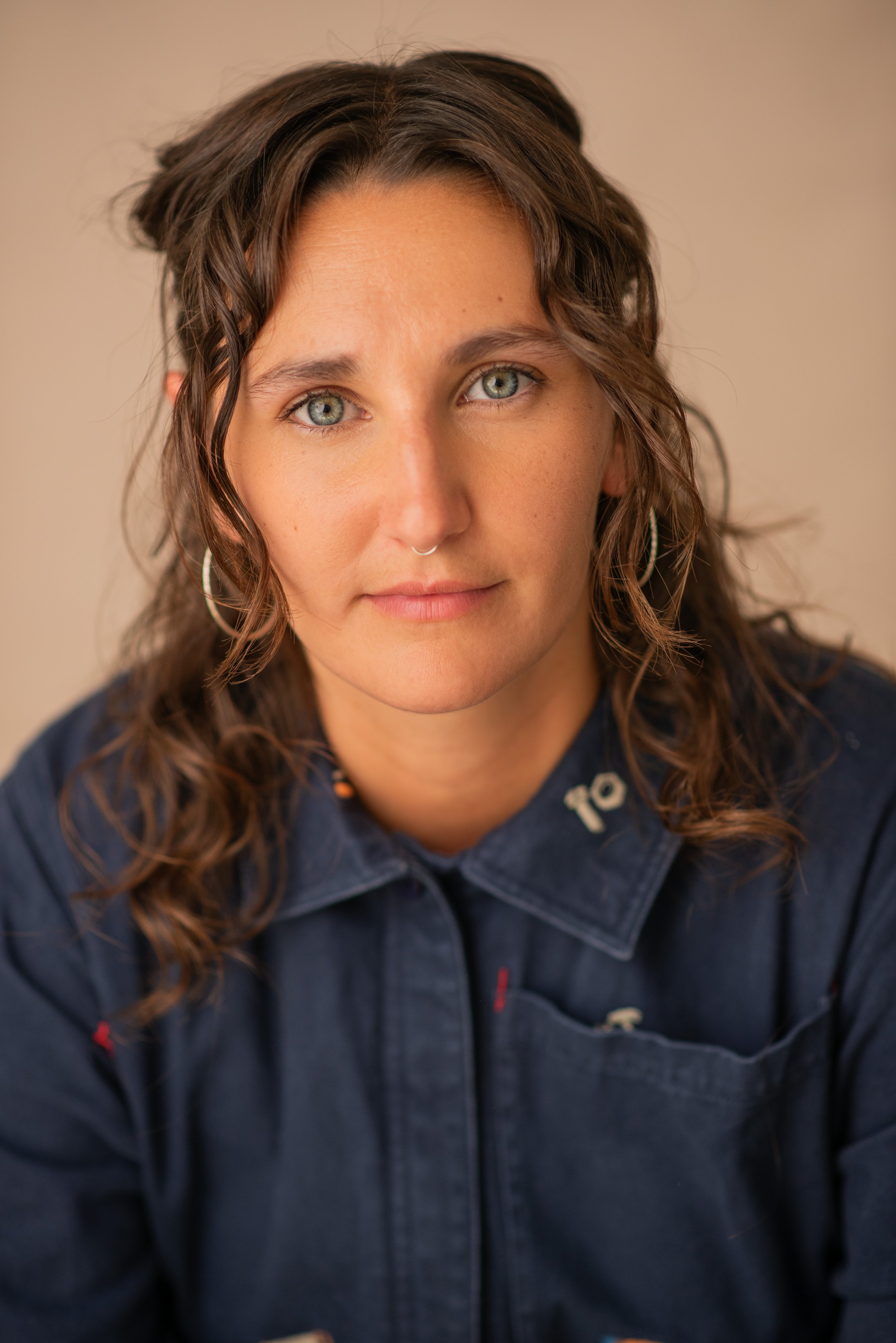Close-up portrait of a woman with wavy brown hair, fair skin, and piercing blue eyes, wearing hoop earrings and a dark denim shirt.