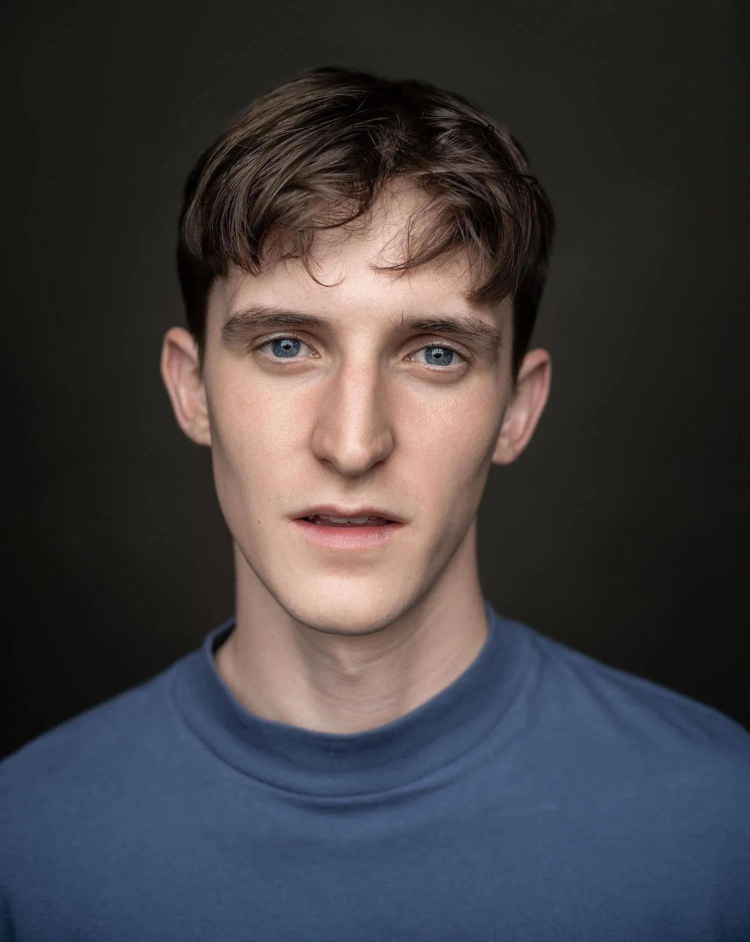 A young man with blue eyes and brown hair, wearing a blue shirt, facing the camera with a neutral expression against a dark background.