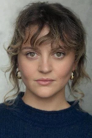 A woman with shoulder-length wavy hair, wearing hoop earrings and a navy blue top, looking directly at the camera.