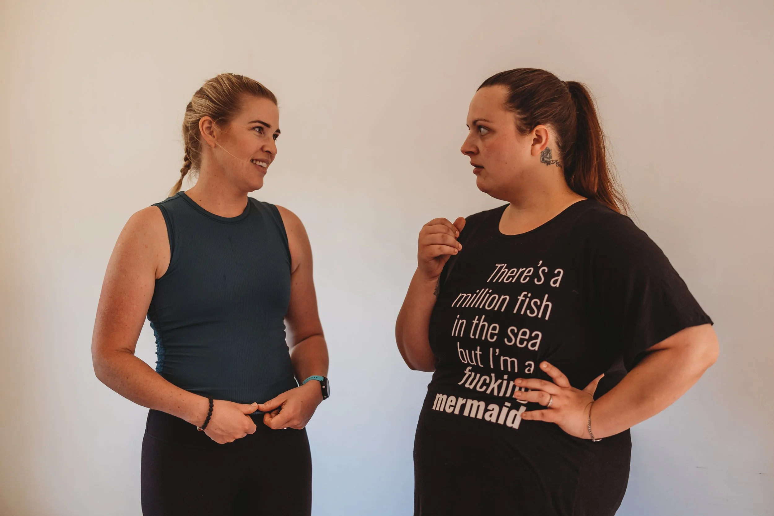 Two women talking, one smiling and the other with a serious expression, against a plain wall.
