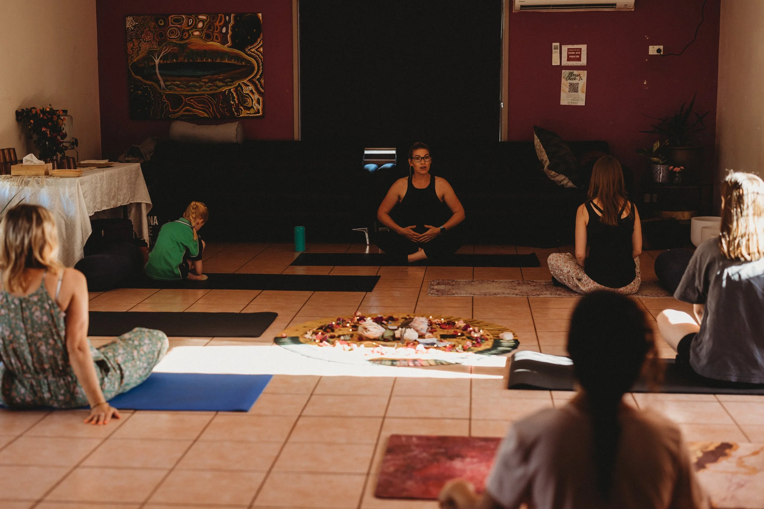 A group of people participating in a yoga or meditation session in a dimly lit indoor space with a woman leading the session, sitting cross-legged with hands on her stomach, surrounded by Yoga mats and a decorative circle of candles and stones on the floor.