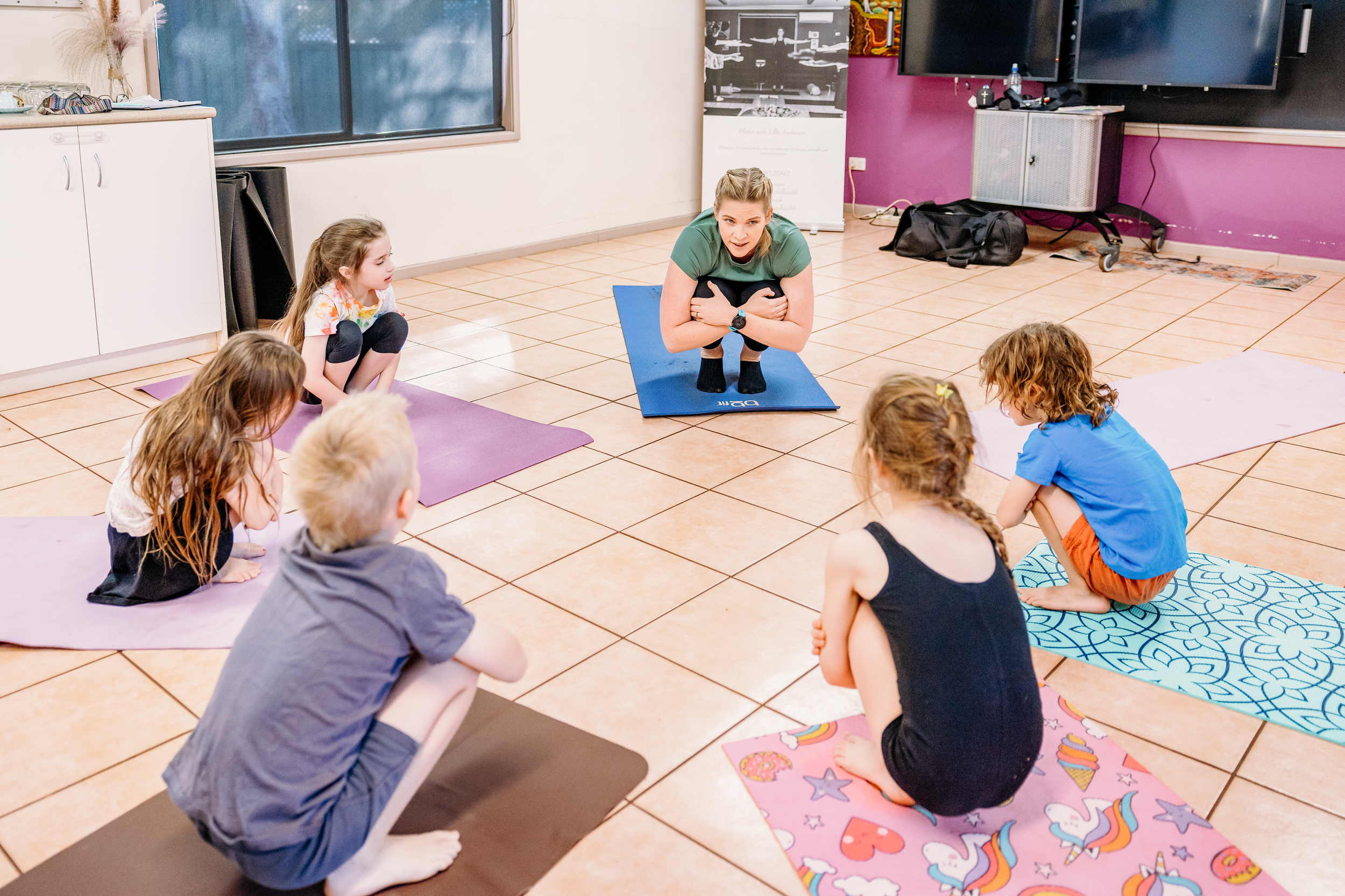 A woman leading a yoga class for children in a room with tiled floors and pink walls, with six children sitting on colorful yoga mats, listening attentively.