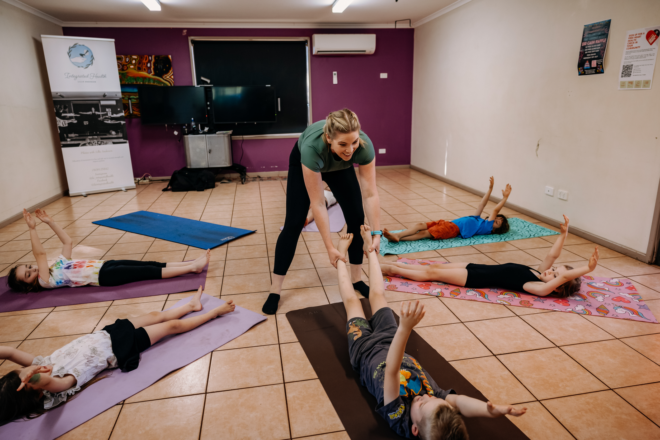 A yoga instructor assisting children in doing yoga poses in a classroom with tiled floor and purple accent wall.